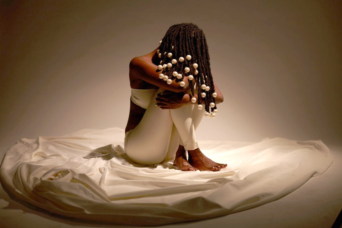 Kelela sits on white fabric wearing beaded hair accessories and a cream-colored outfit against a neutral background.