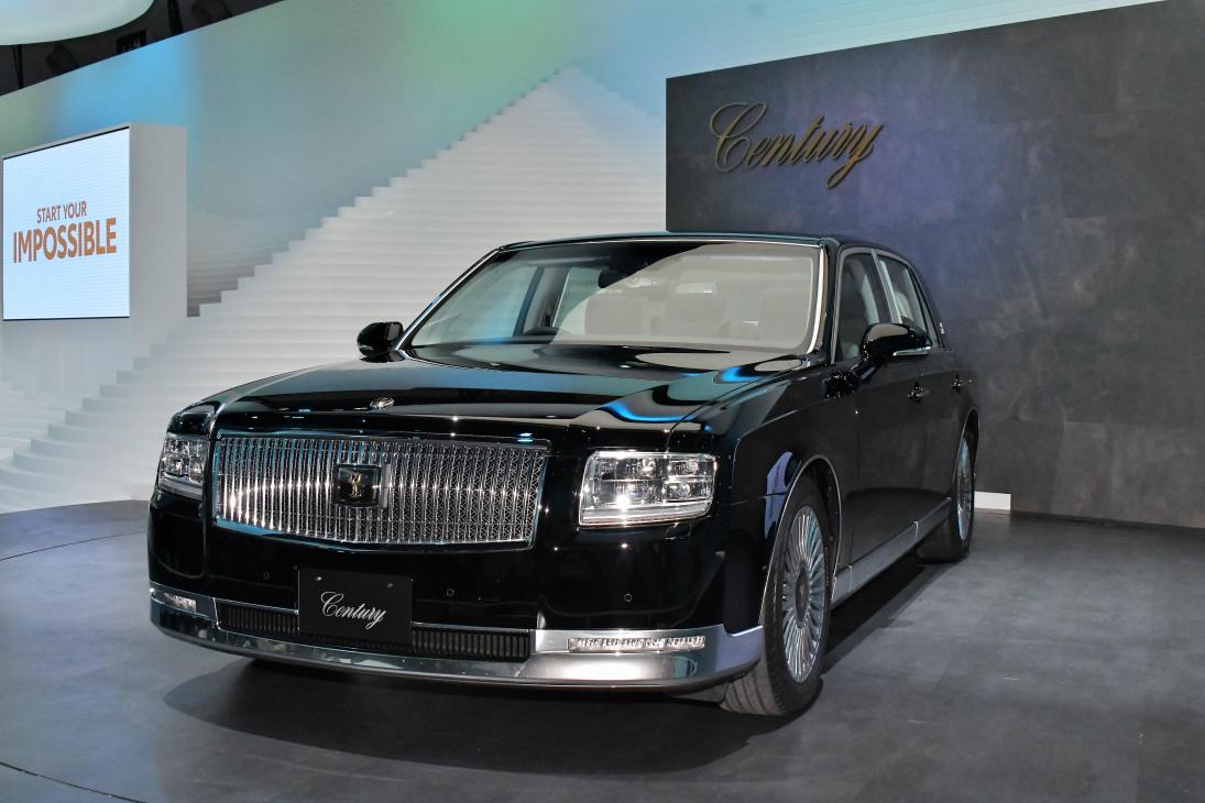 Black Toyota Century luxury sedan displayed at an event with modern architecture in background.