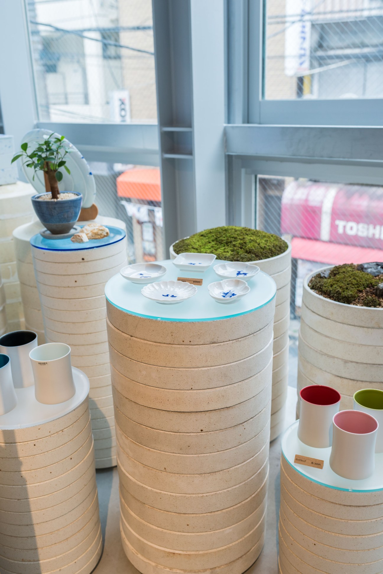 Kihara ceramics display with stacked wooden shelving, moss-topped vessels, and white porcelain cups with blue designs.