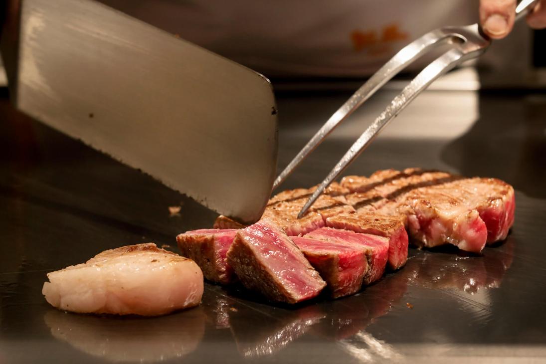Chef using metal utensils to plate sliced Kobe beef on a dark surface.