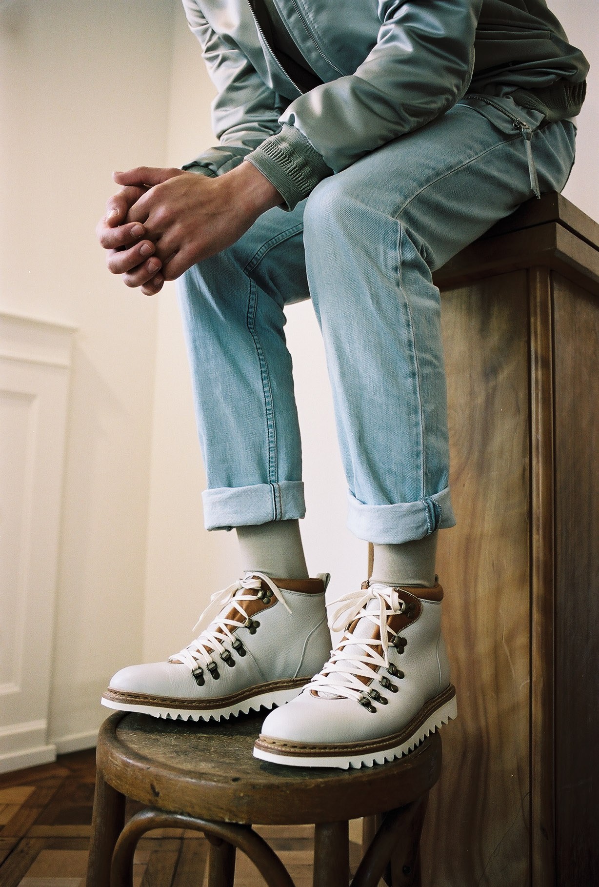 Person wearing white leather hiking boots with brown accents and rolled-up jeans, seated on a wooden stool.
