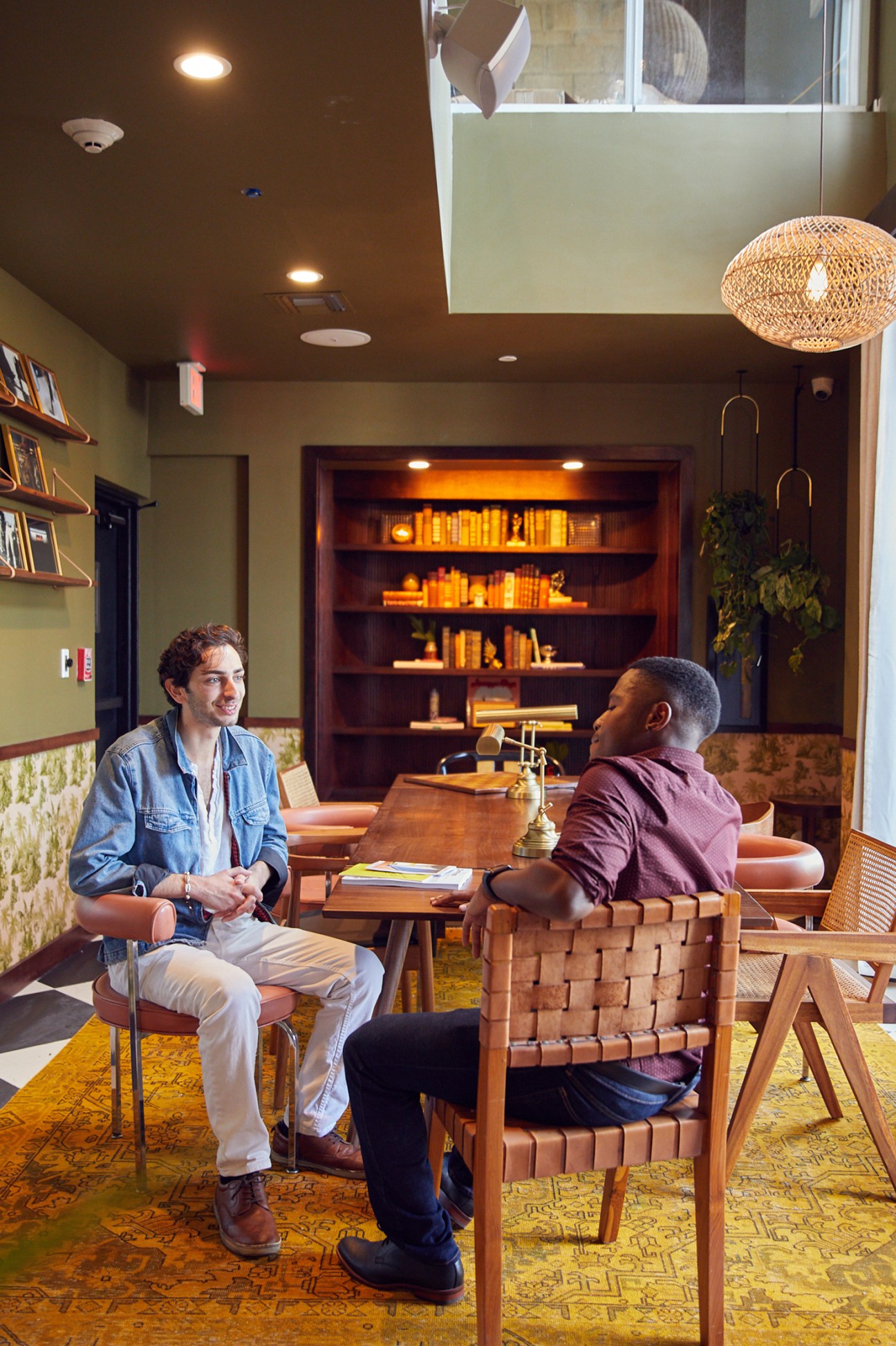 Two men in conversation at a wooden table in a modern hotel lounge with bookshelves and woven pendant light.