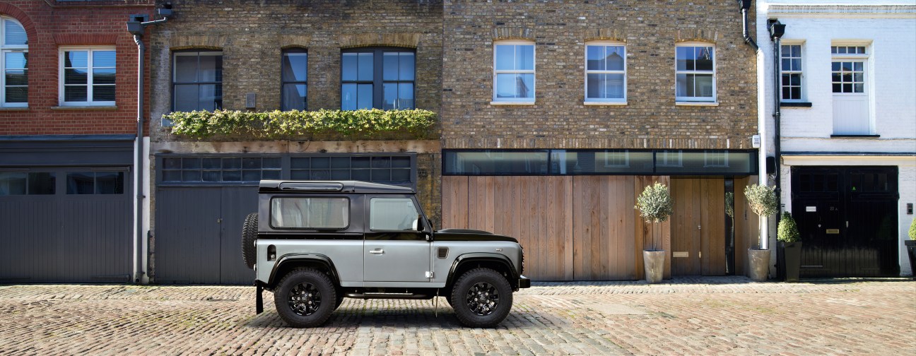 Silver Land Rover Defender parked on cobblestone street in front of brick buildings.
