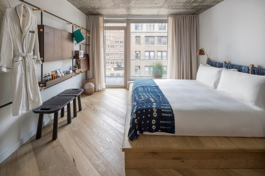 Modern hotel bedroom at Made Hotel New York with wooden furnishings, blue patterned throw, and city views through floor-to-ceiling windows.