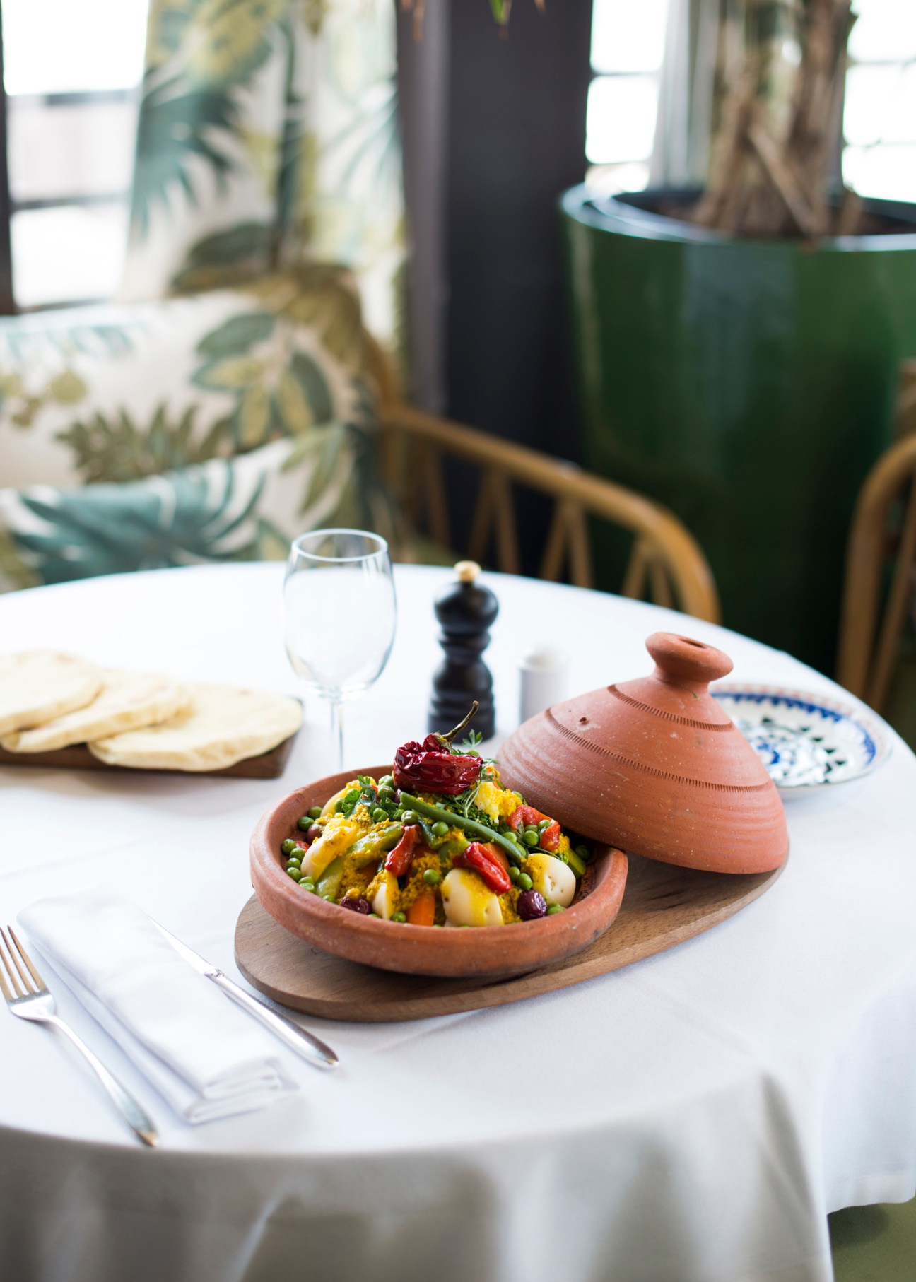 Moroccan tagine dish with vegetables served in terracotta bowl at white-tablecloth restaurant with plants and windows.