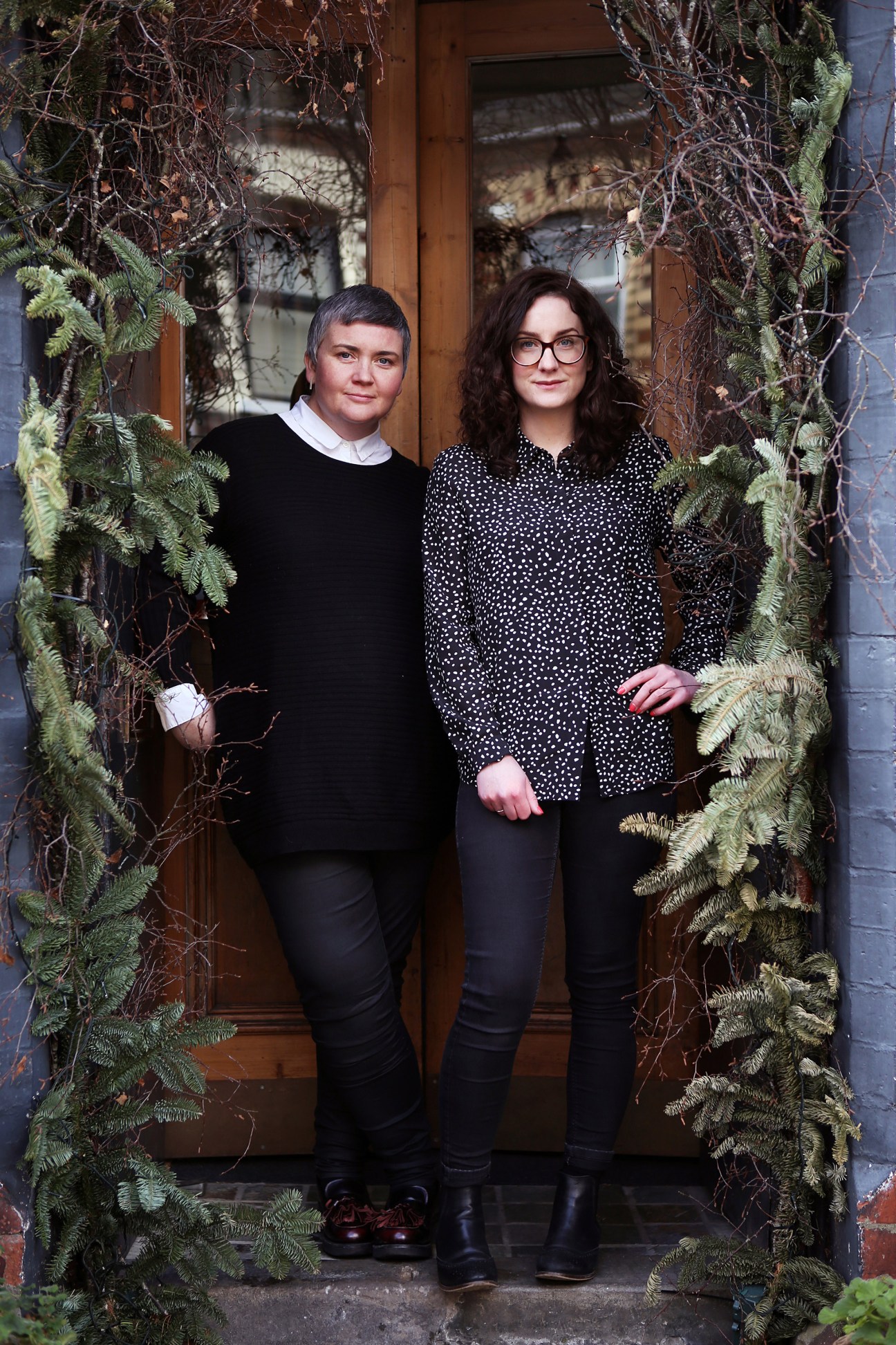 Two women stand in a doorway framed by ivy and climbing plants, smiling at the camera.