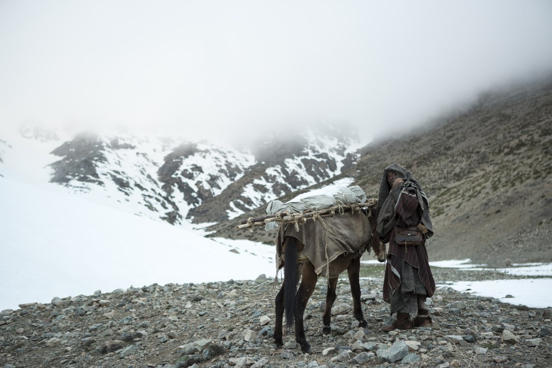 A person in dark robes stands with a donkey loaded with supplies in a mountainous landscape with snow-capped peaks.