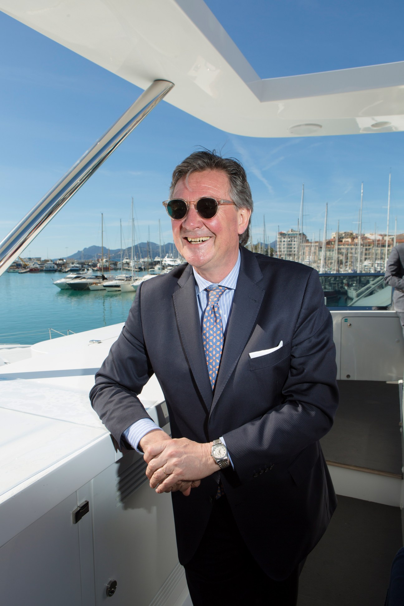 Man in business suit and sunglasses standing on a yacht with Cannes marina in the background.