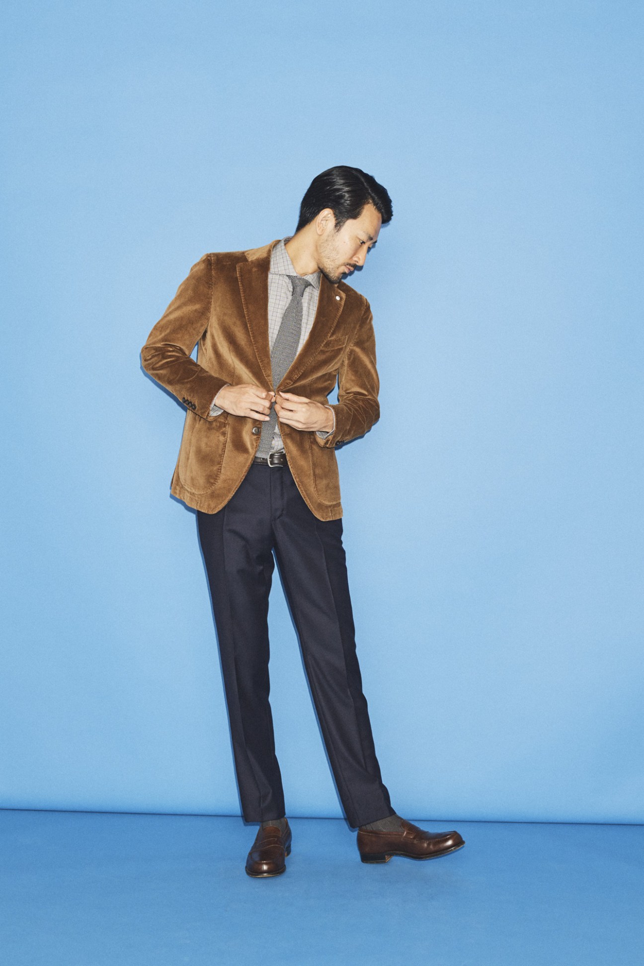 Man wearing a brown velvet blazer and dark trousers against a blue background.