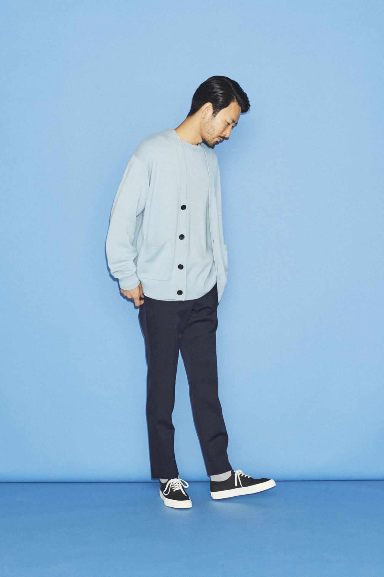 Man wearing light grey cardigan with black buttons and dark trousers against blue background.