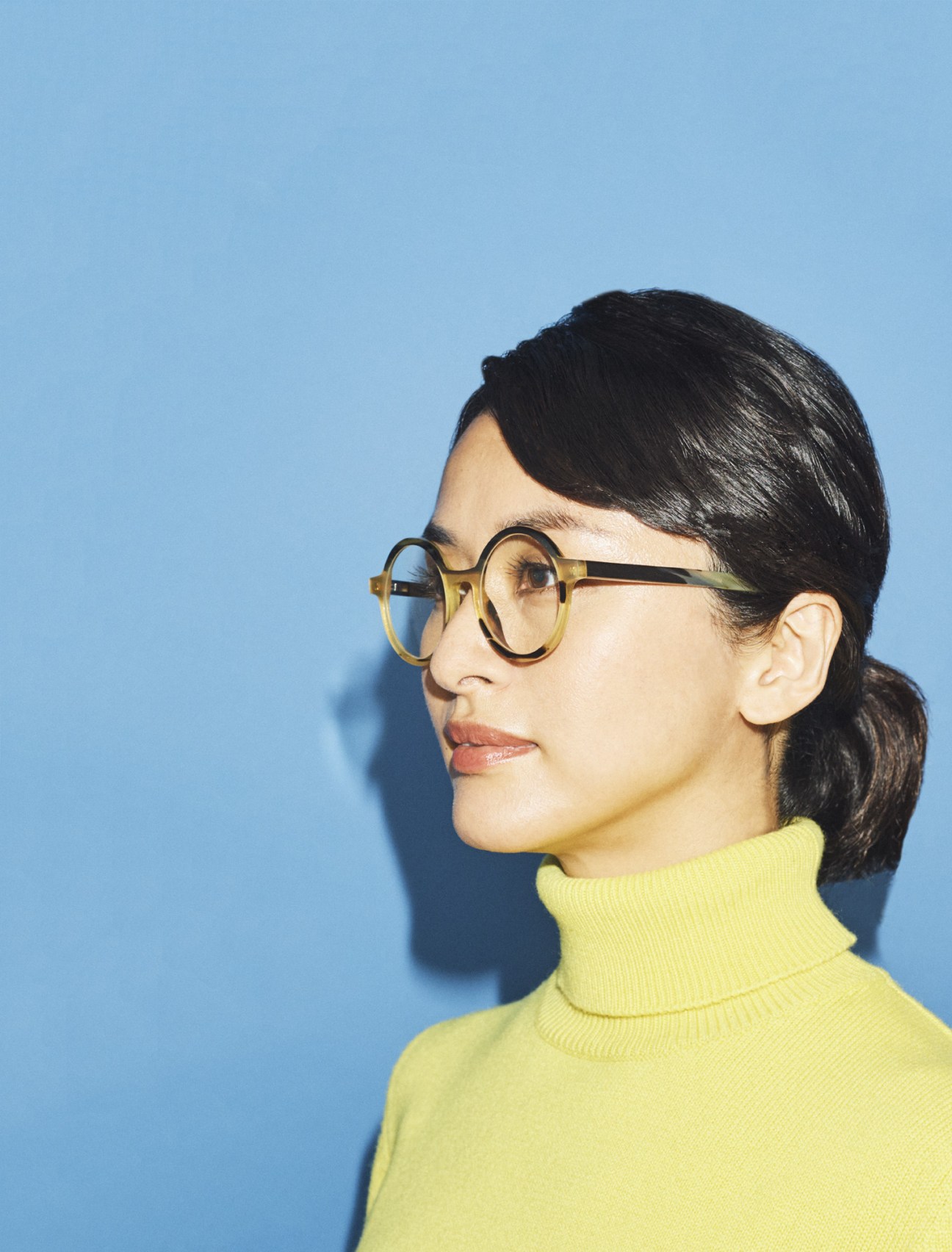 Woman wearing round tortoiseshell glasses and yellow turtleneck against blue background.