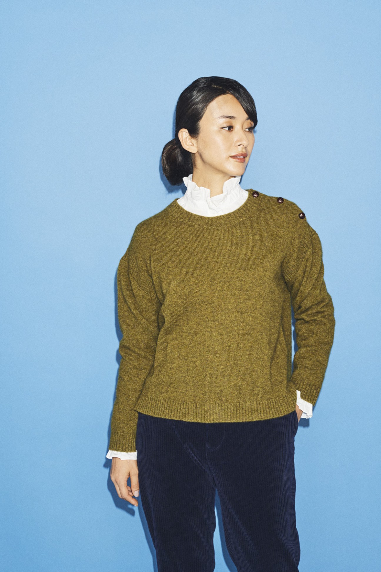 Woman in olive-green knit sweater with white collar shirt against blue background.