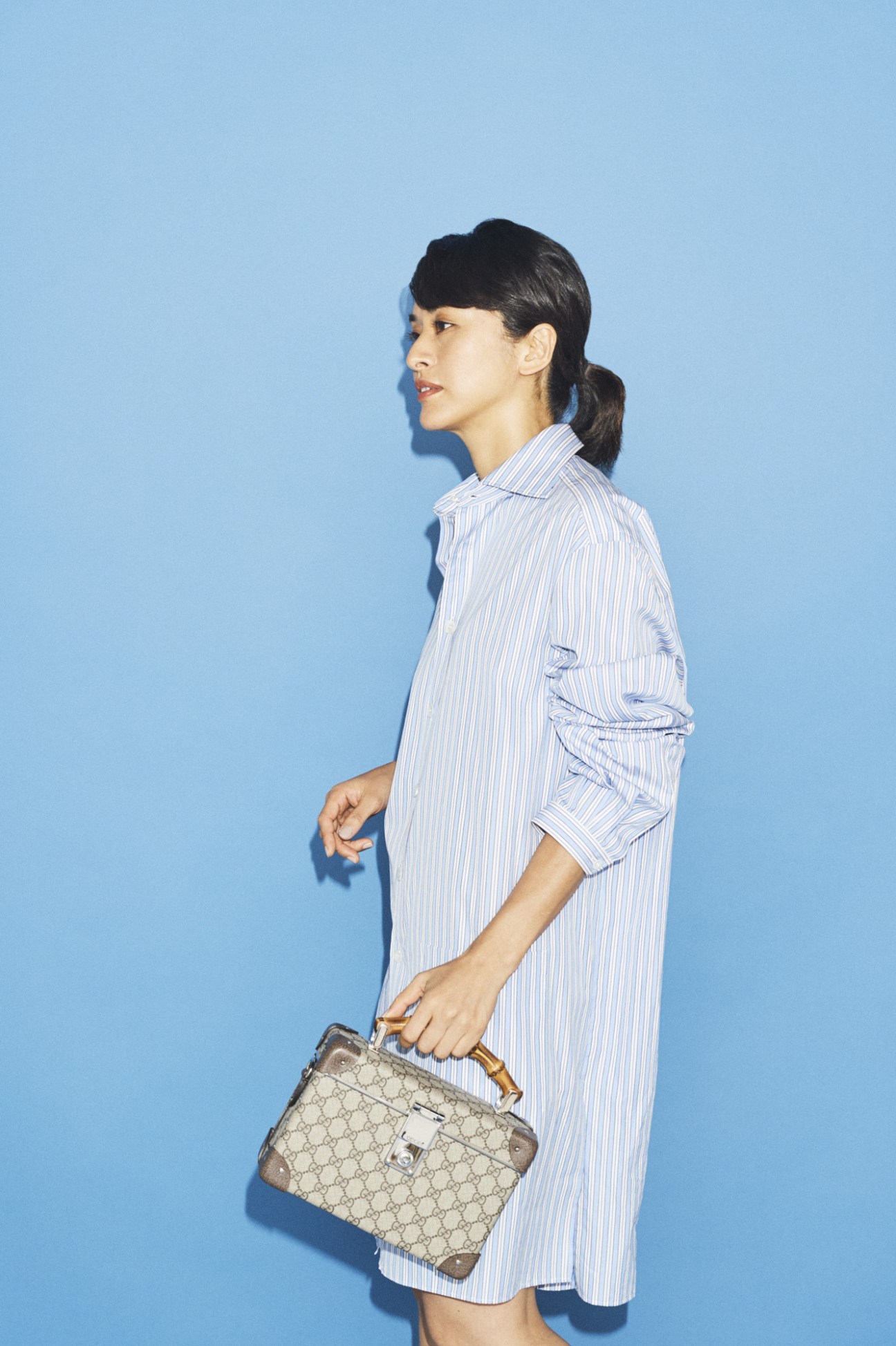 Woman wearing light blue striped shirt dress holding patterned luggage bag against blue background.