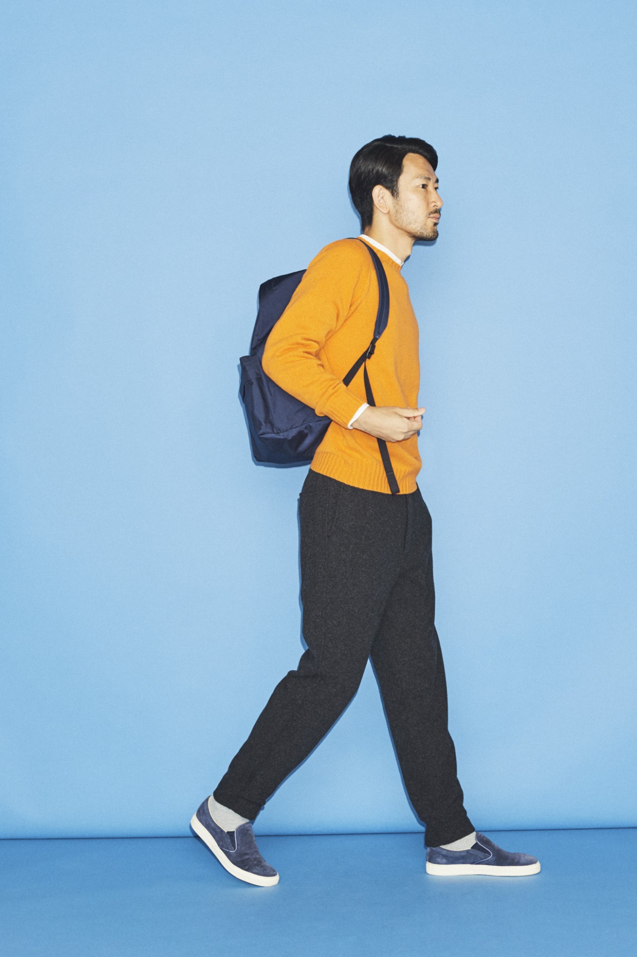 Man wearing a jumper by Morris & Sons, shirt by Adam Et Rope, trousers by Comoli, socks by Uniqlo, trainers by Yoak, and carrying a backpack by Aeta.
