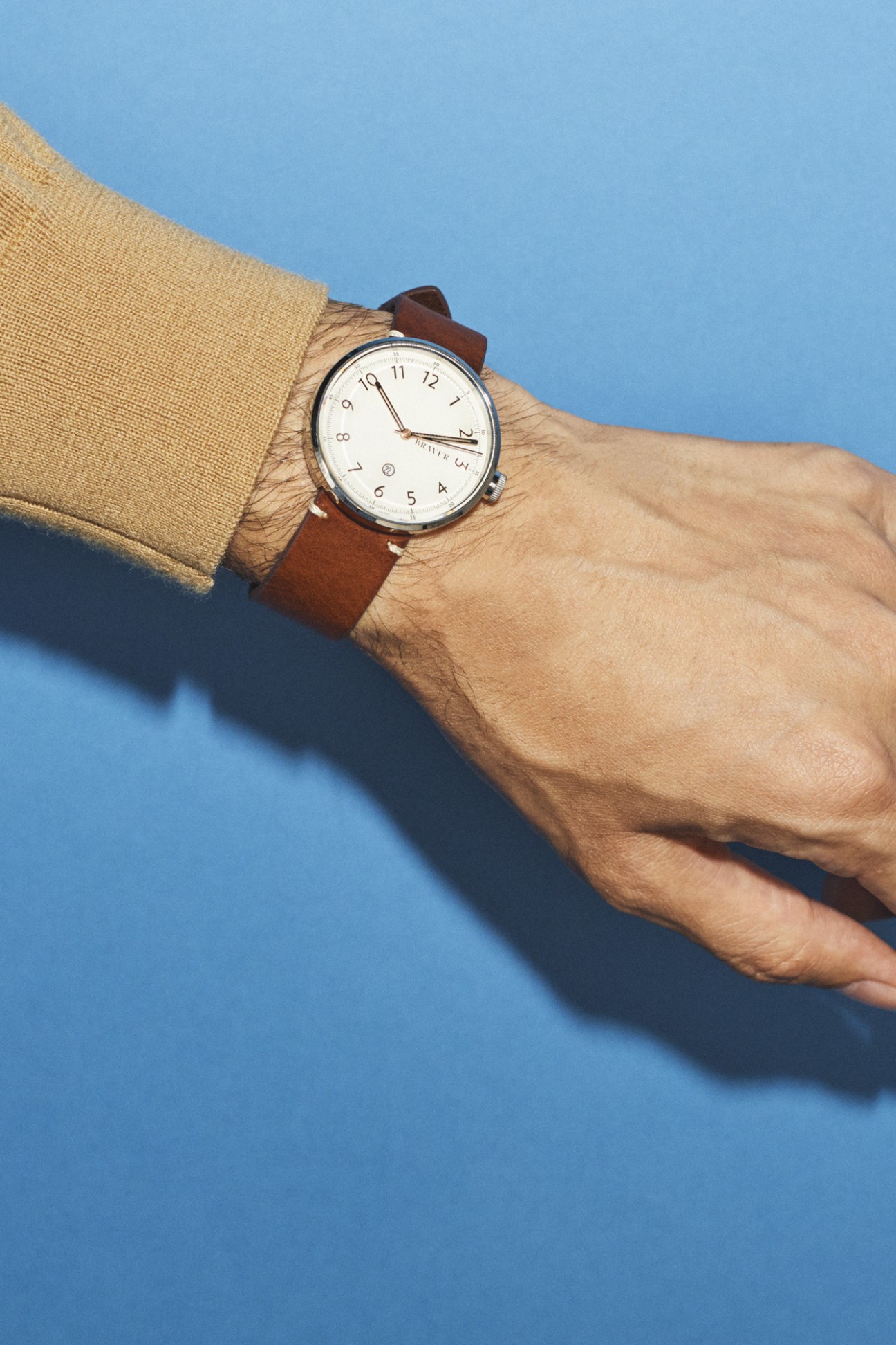Man's wrist wearing a Bravur watch with white dial and brown leather strap against blue background.