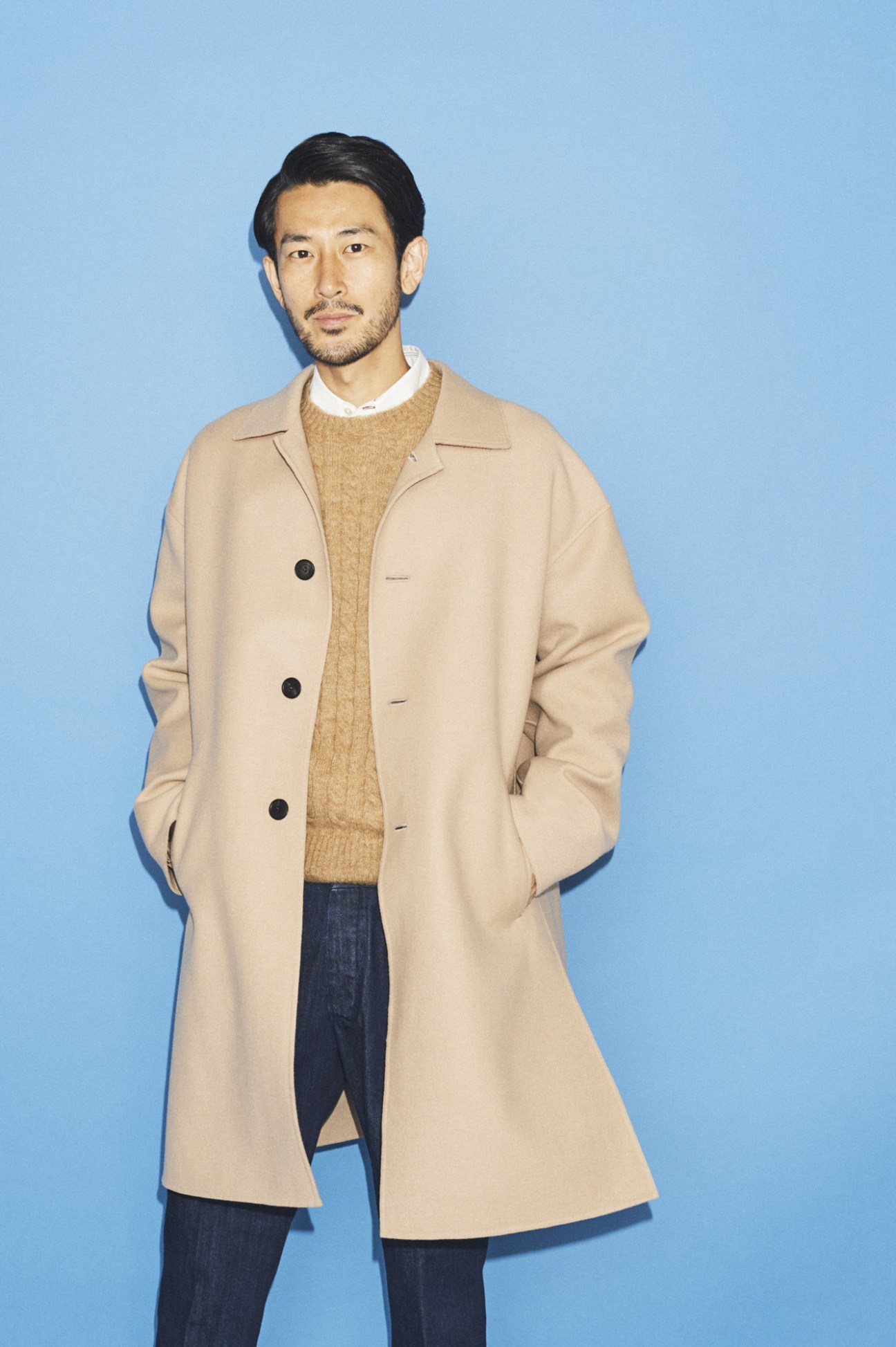 Man wearing beige coat, camel knit sweater and dark jeans against blue background