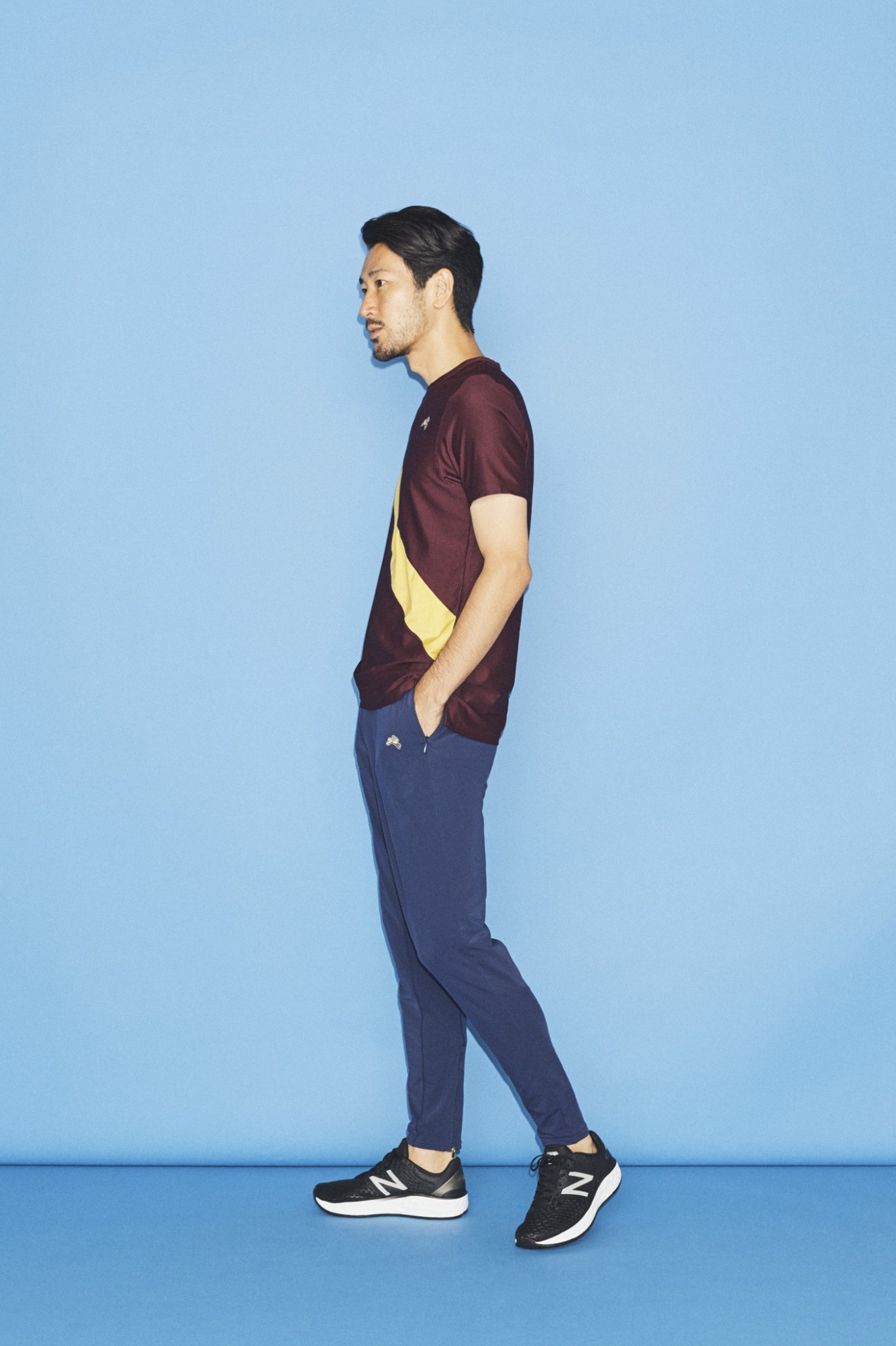 Man in profile wearing burgundy and yellow colorblock t-shirt, navy trousers, and black New Balance sneakers against blue background.