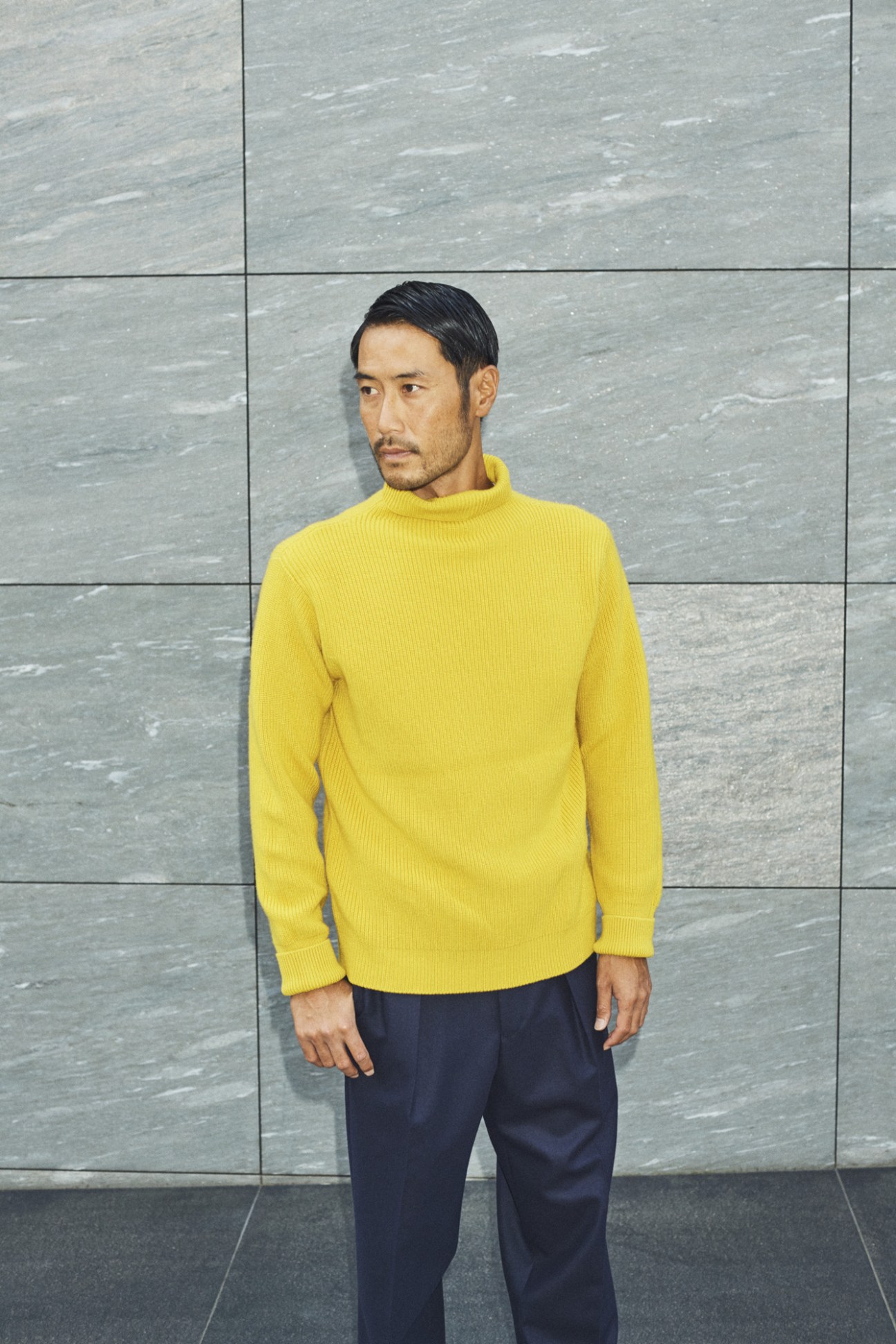 Man wearing yellow ribbed turtleneck sweater and navy trousers against grey tiled wall