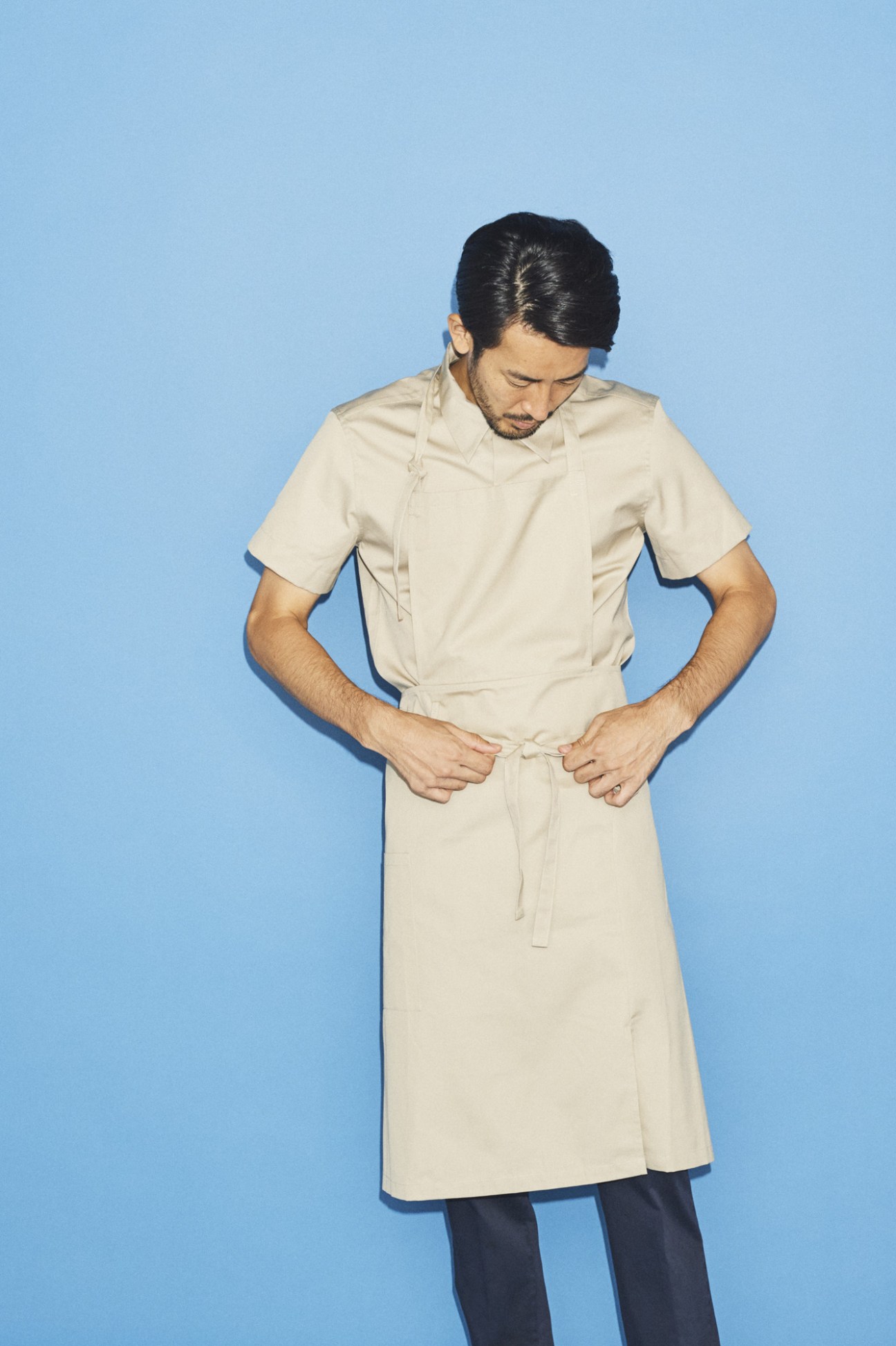 Man in cream button-up shirt and light-colored skirt-like garment tied at waist against blue background.