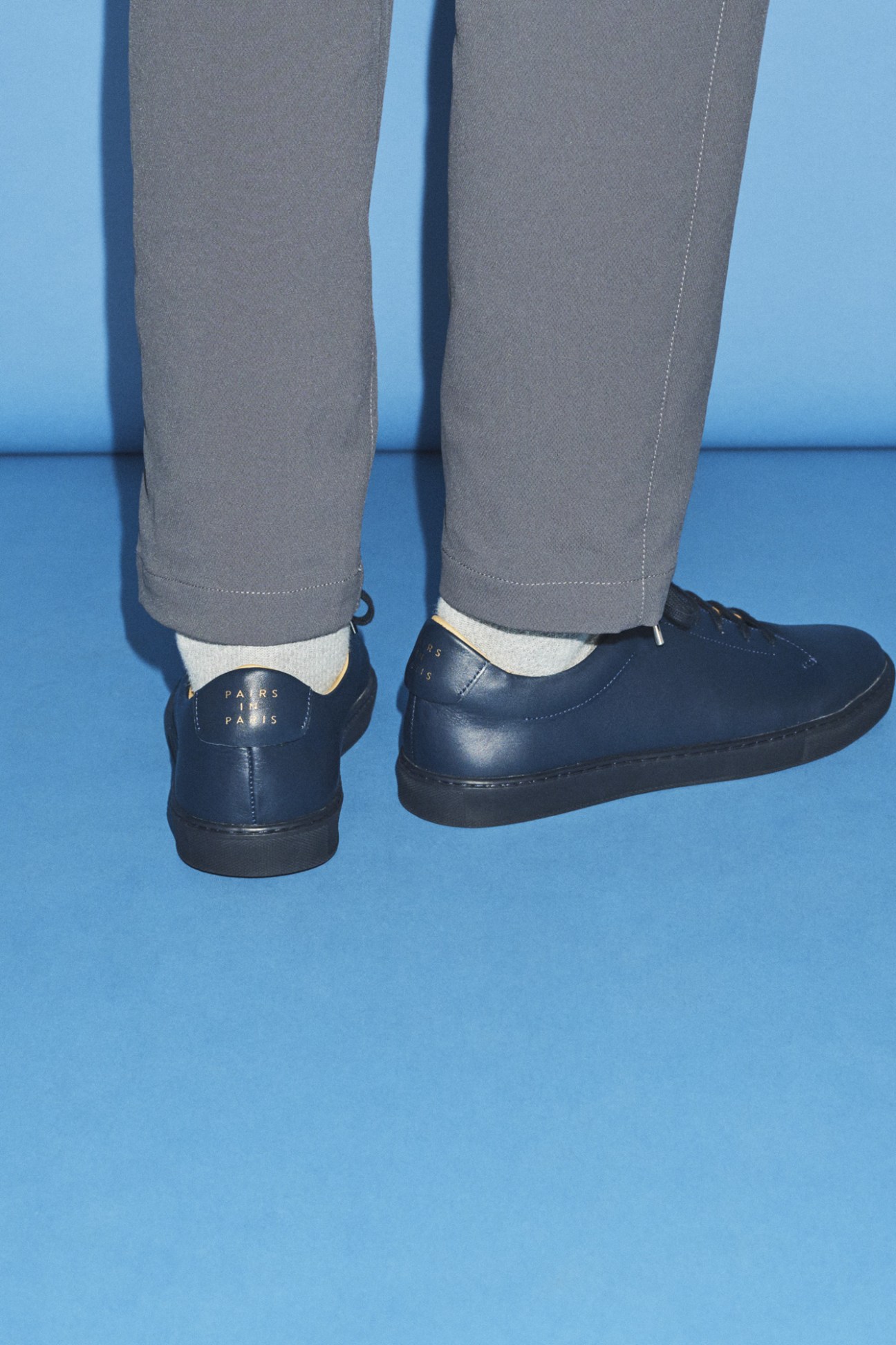 Person wearing grey trousers, white socks and navy Pairs in Paris trainers against blue background
