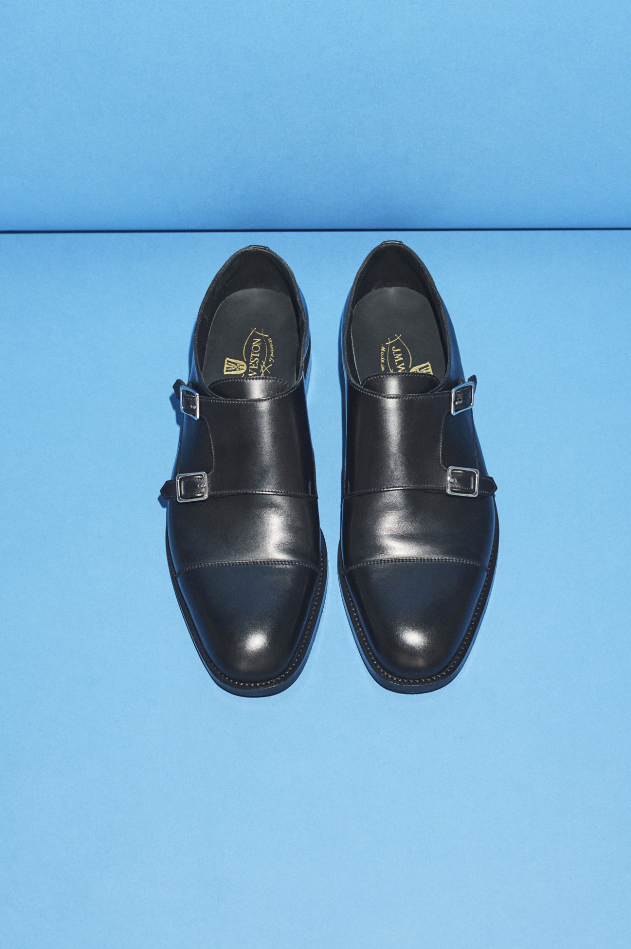 Pair of black leather monk strap shoes with metal buckles on a light blue background.