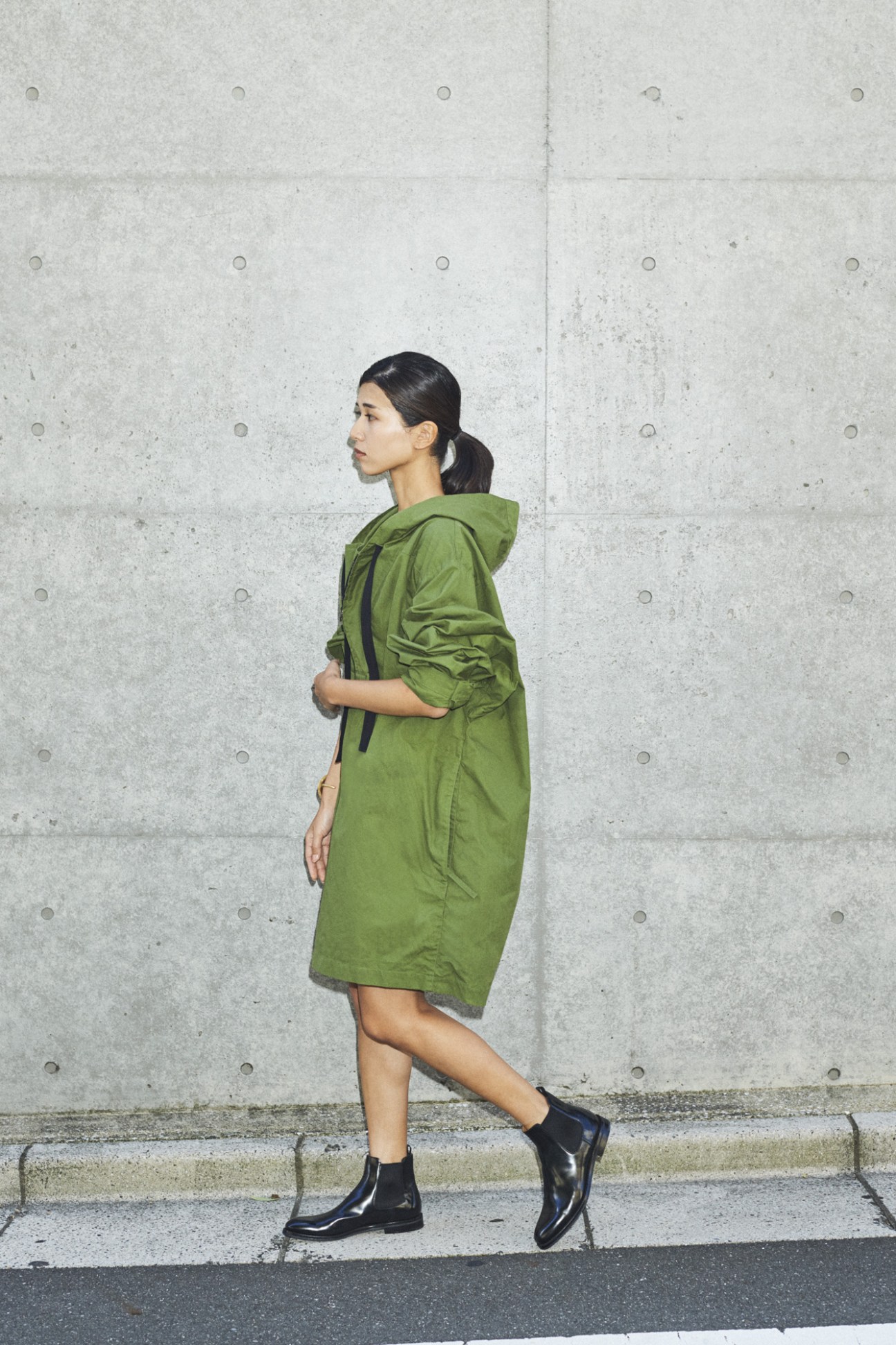 Woman in profile wearing a green hooded jacket against a concrete wall.