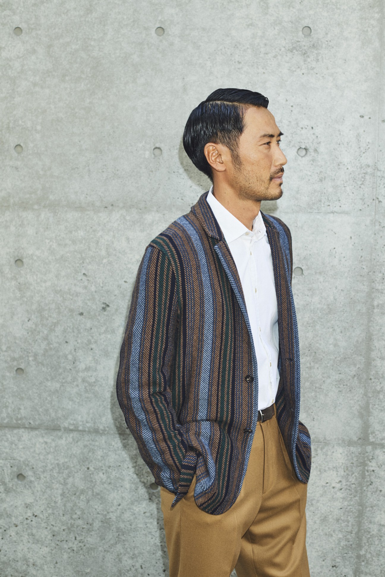 Man in profile wearing a striped navy and brown cardigan over a white shirt and tan trousers against a concrete wall.