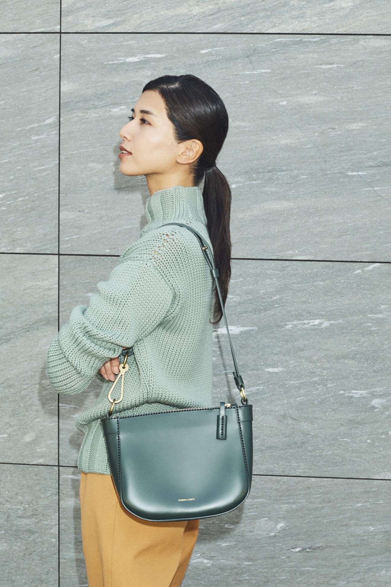 Woman in sage-green knit sweater and mustard trousers carrying a green leather shoulder bag against a textured grey wall.