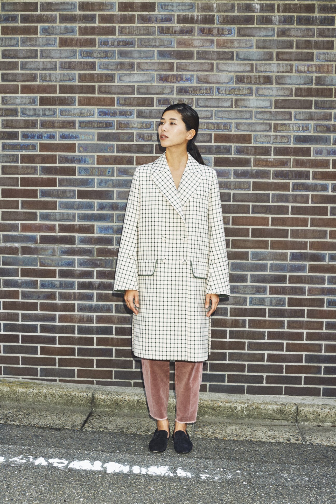 Woman in white checkered blazer and pink trousers standing against brick wall