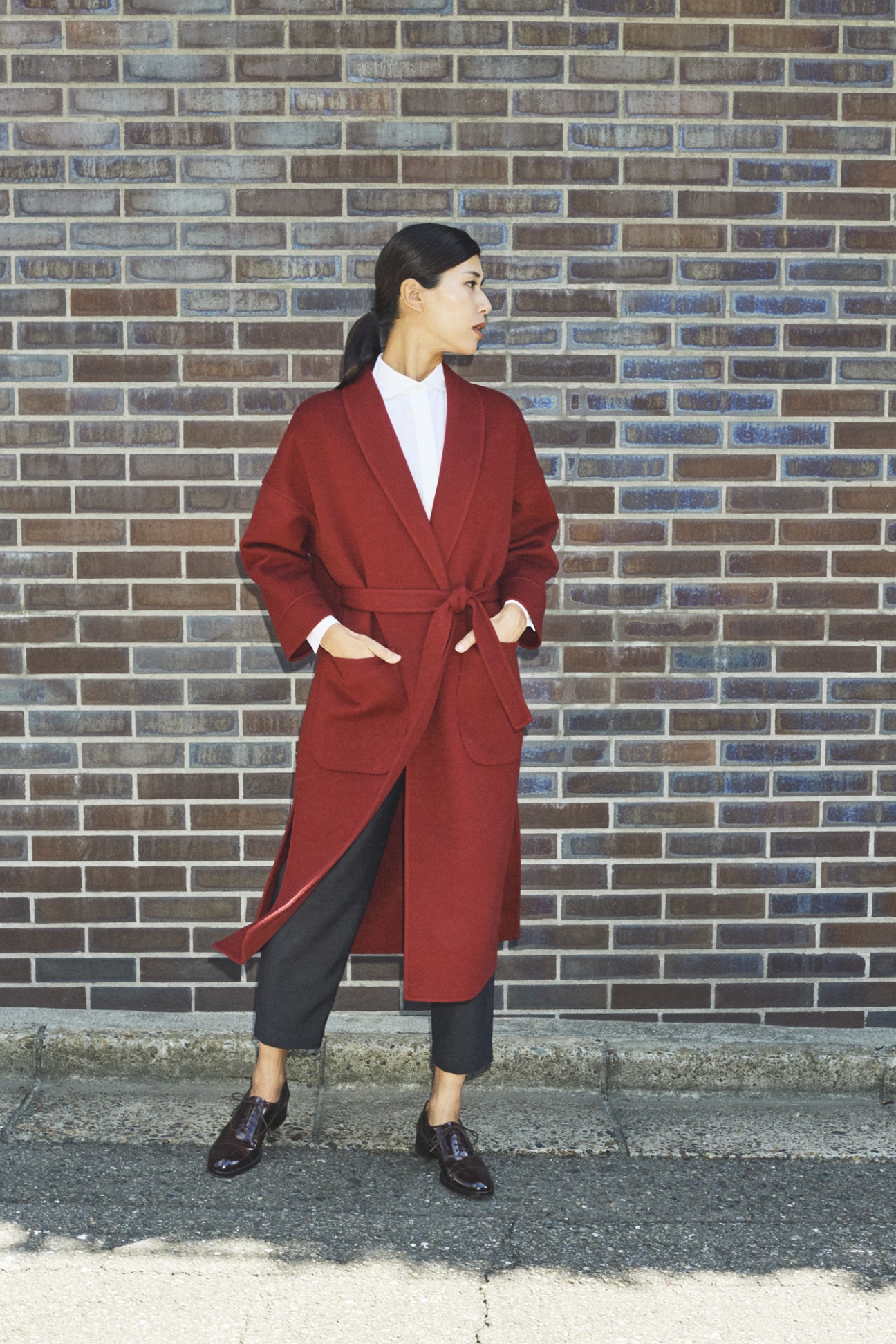 Woman in red wool coat and white shirt standing against brick wall.