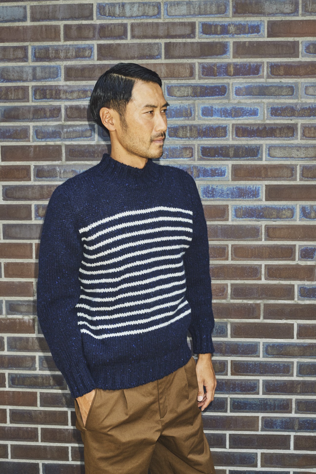 Man wearing navy striped sweater with white horizontal stripes and tan trousers against brick wall.