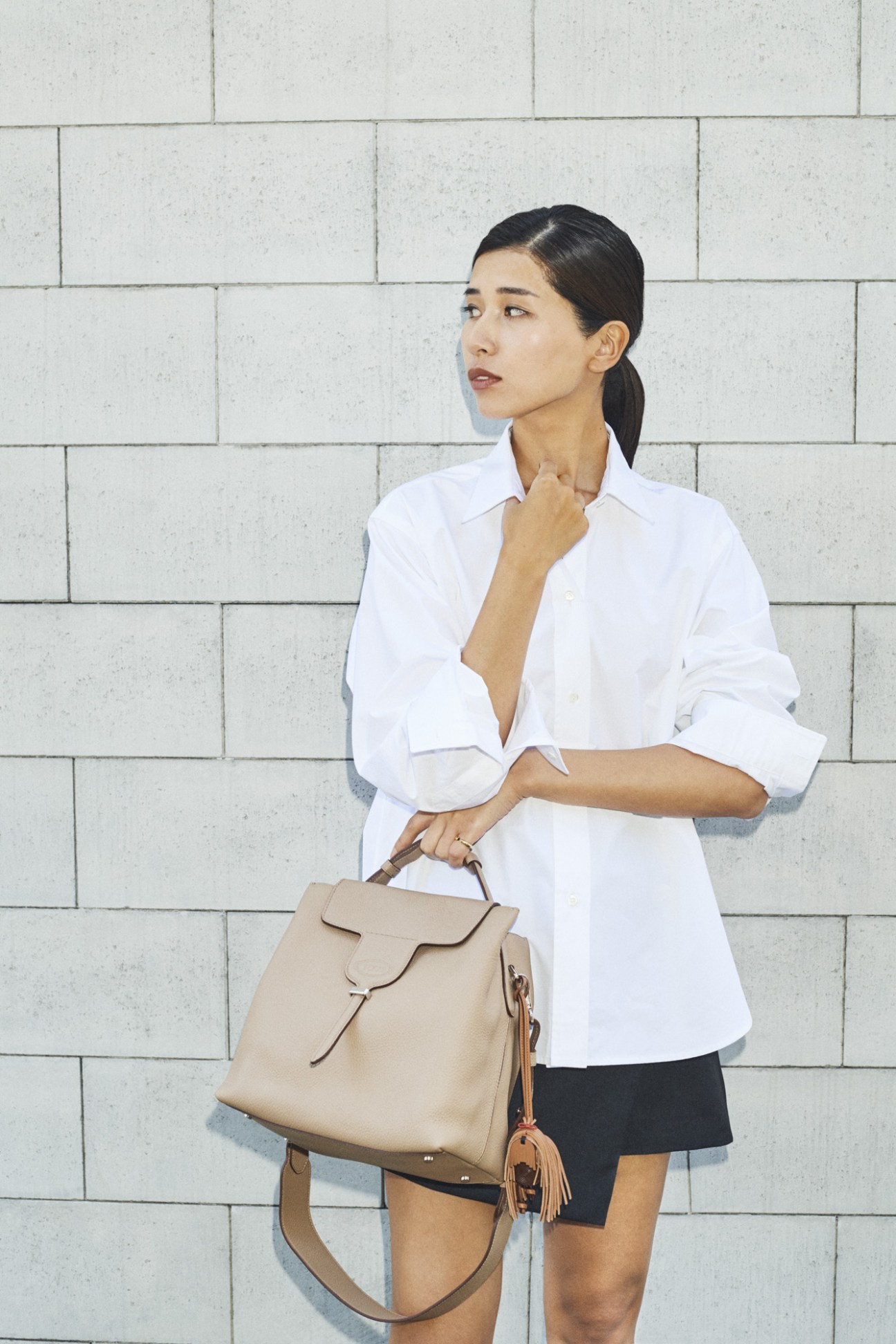 Woman in white shirt holding beige leather handbag against brick wall.