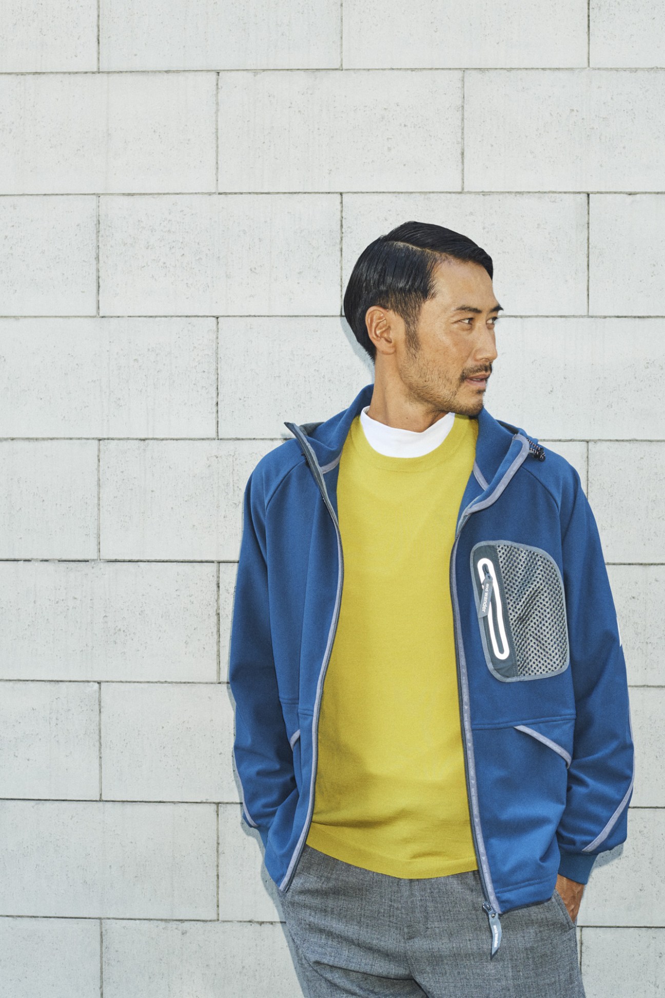 Man wearing blue zip-up jacket with mesh pocket, yellow t-shirt, and grey trousers against white brick wall.