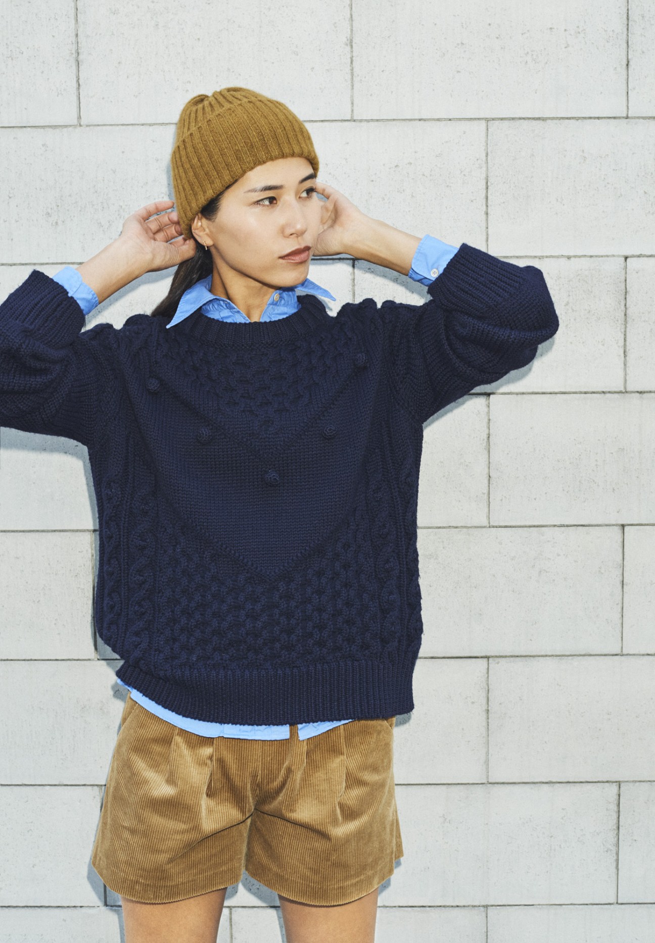 Woman wearing navy cable-knit sweater, light blue shirt, tan shorts, and mustard beanie against white tiled wall.