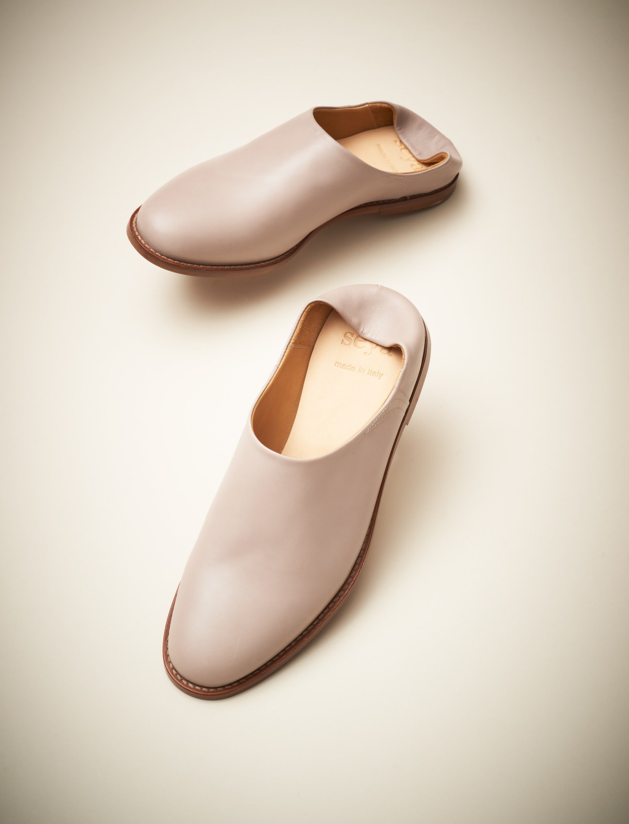 Two cream leather babouche slippers with brown trim and open heels against a neutral background.