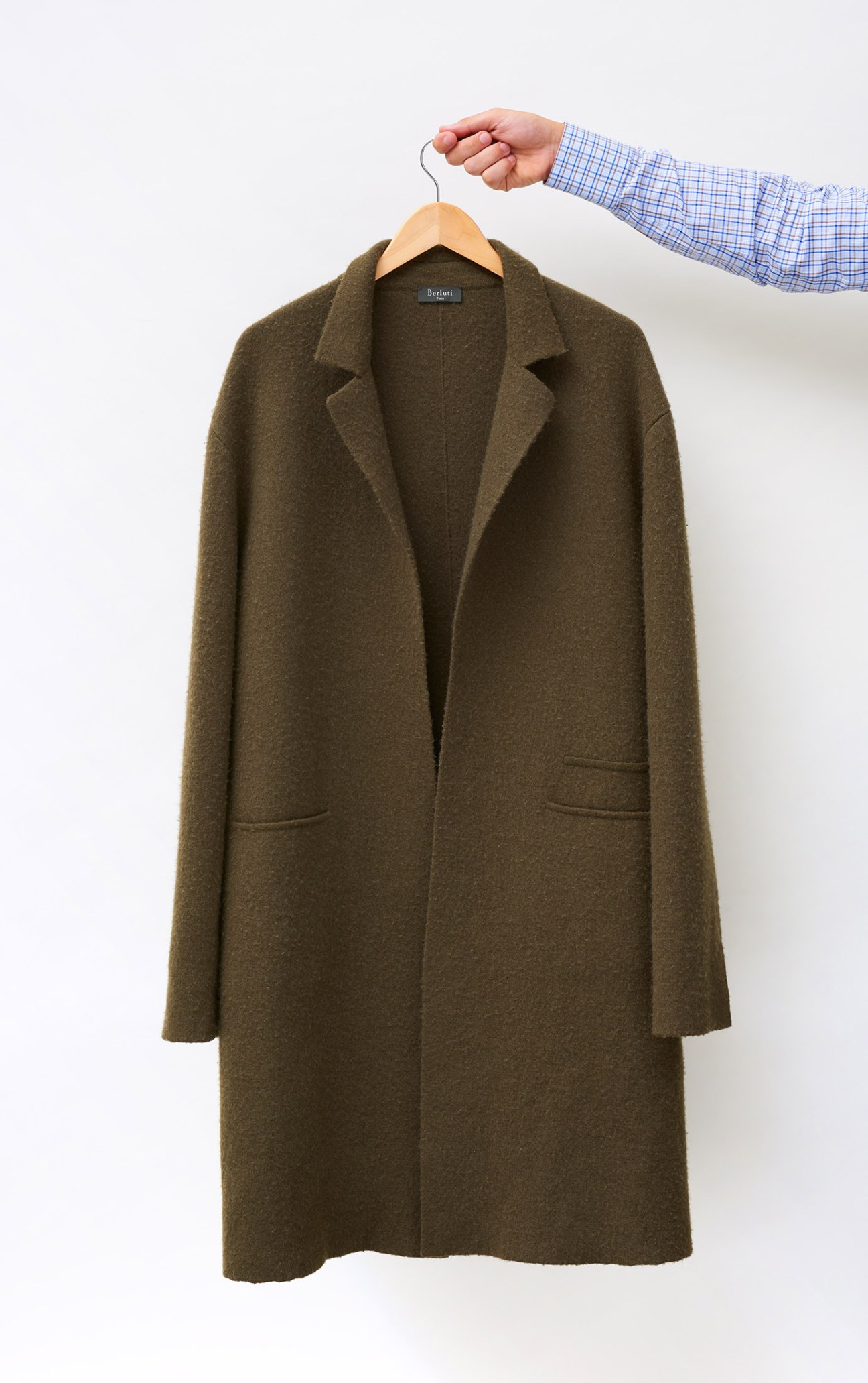 Oversized olive-green virgin wool coat by Berluti held on wooden hanger against white background.