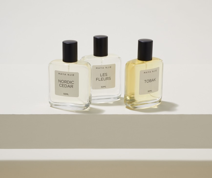 Three Maya Njie perfume bottles with clear and amber liquid and black caps displaying Nordic Cedar, Les Fleurs, and Tobak fragrances.
