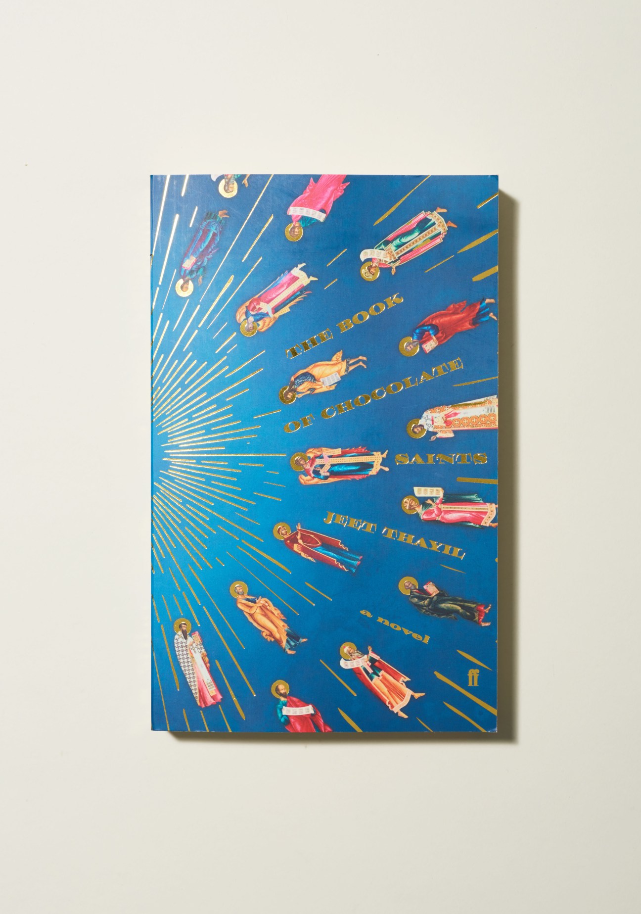 Book cover of The Book of Chocolate Saints by Jeet Thayil, featuring illustrated figures with halos on a blue background with gold radiating lines.