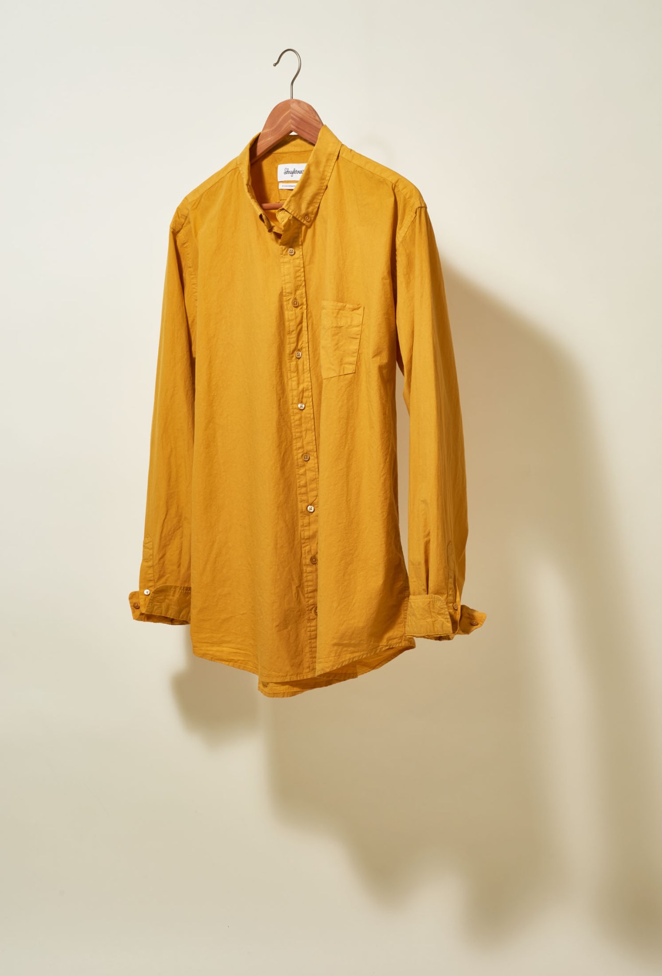 Gold mustard-colored button-up shirt on wooden hanger against cream background.