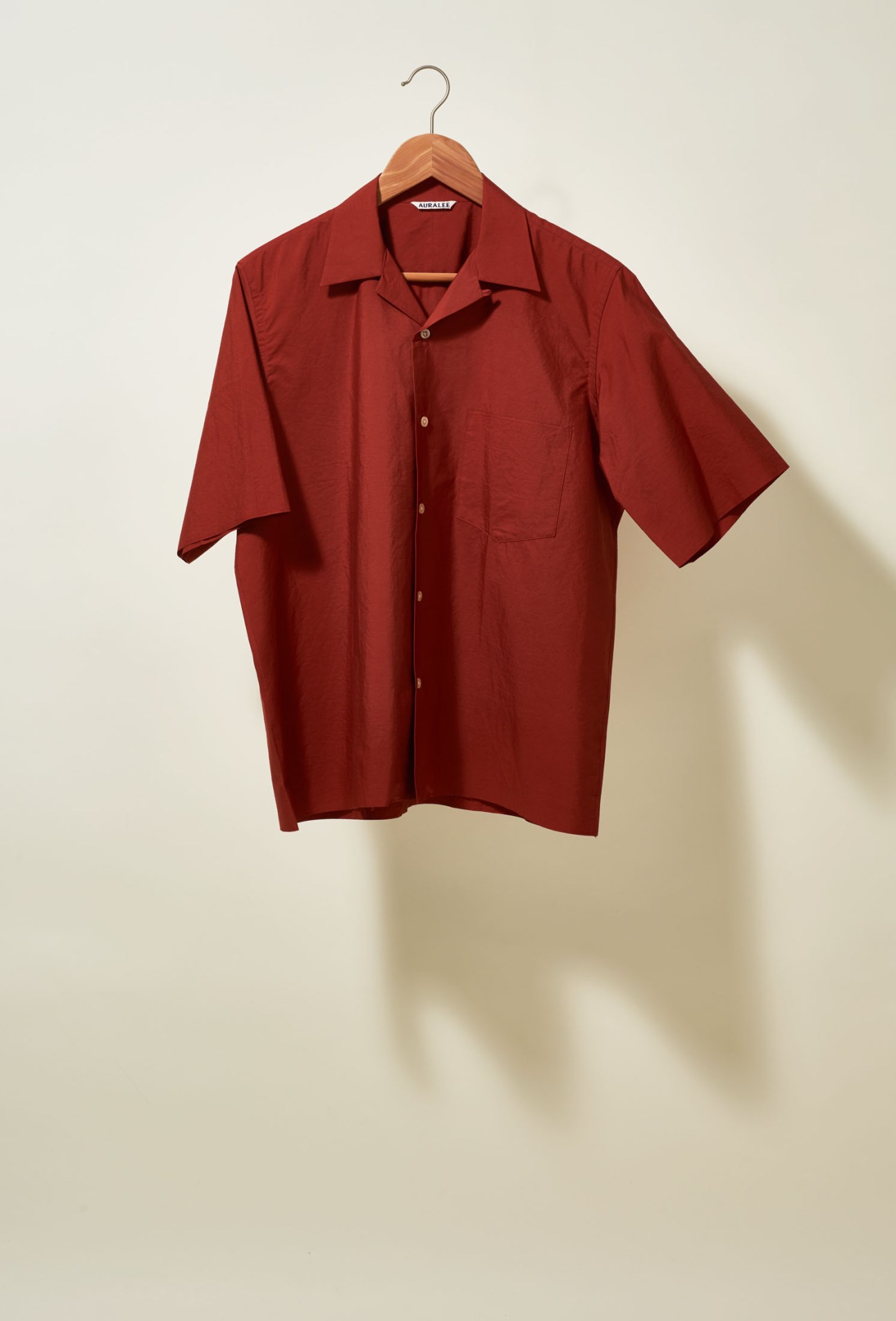 Brick-red camp-collar short-sleeved shirt by Auralee on wooden hanger against neutral background.