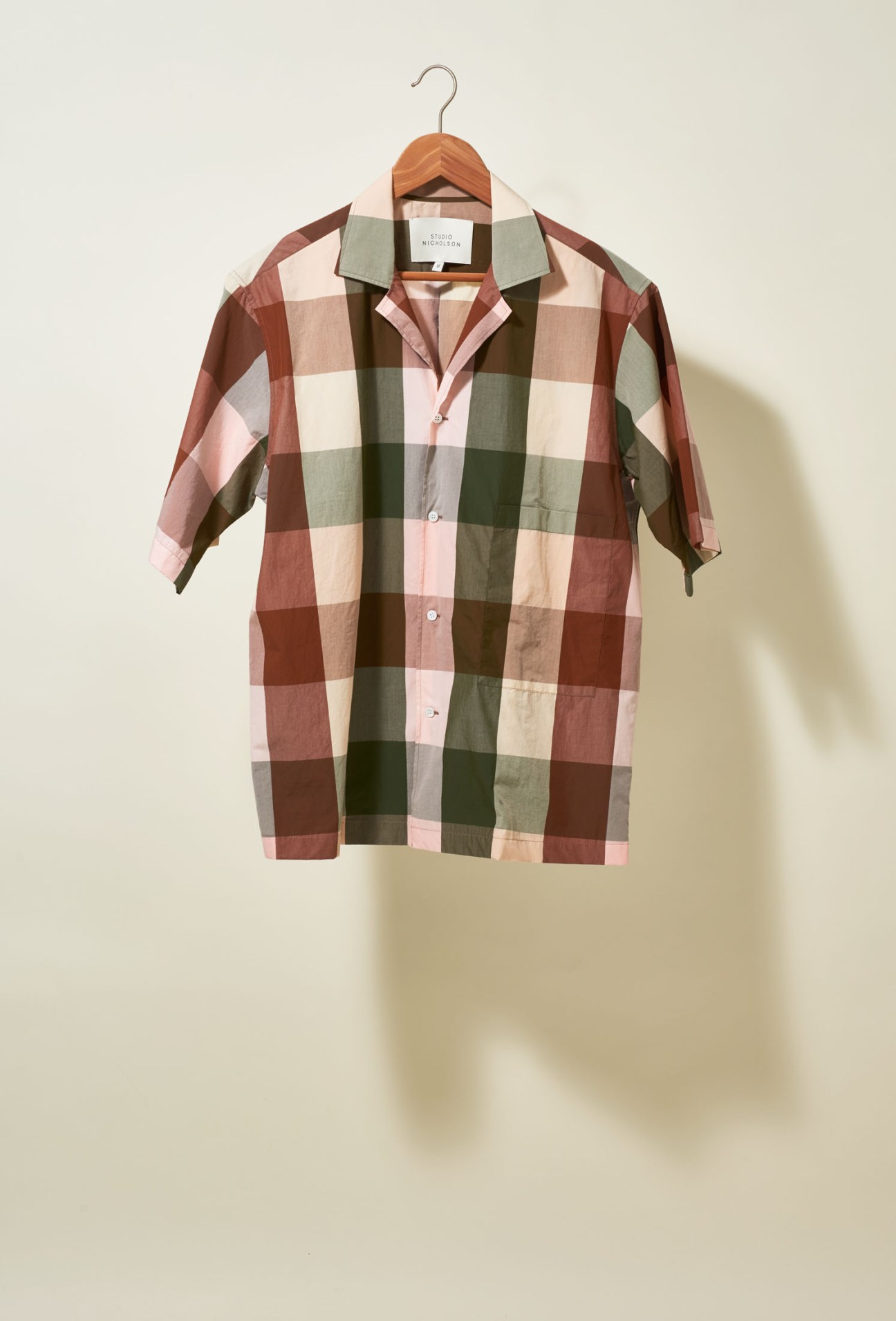 Studio Nicholson Madras-checked boxy shirt in burgundy, cream and olive green on wooden hanger.