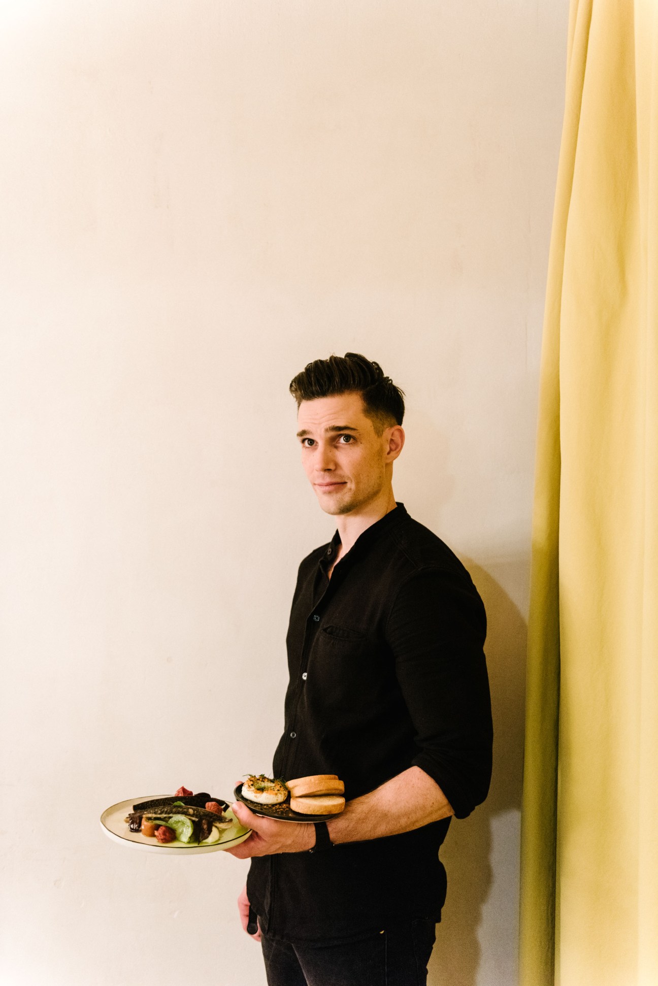 A man in a black shirt holds two plates of food while standing against a white wall with a yellow curtain.