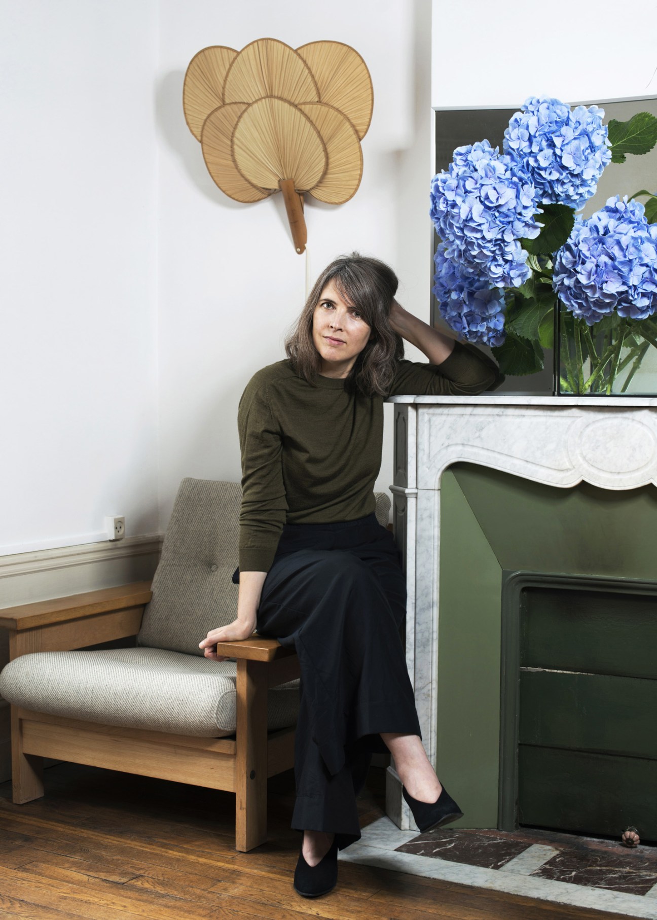 Clarisse Demory, interior designer, sitting on a wooden chair in a bright room with a decorative fan and blue hydrangeas on mantelpiece.