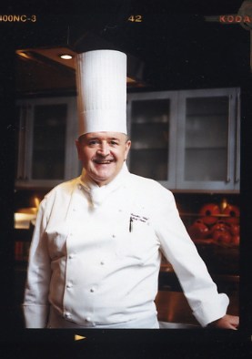Chef in white uniform and tall white chef's hat smiling at camera in restaurant kitchen.
