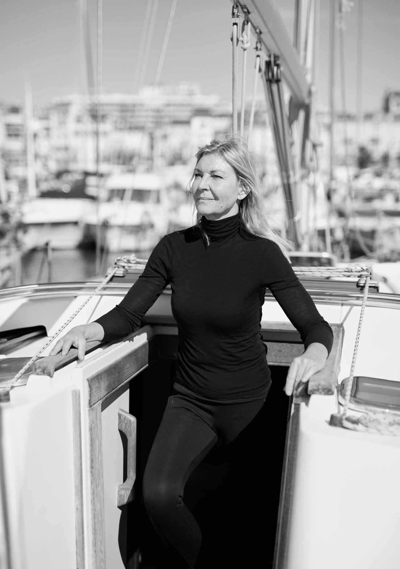 Woman in black turtleneck standing on a sailboat in Cannes harbour with coastal buildings blurred in background.
