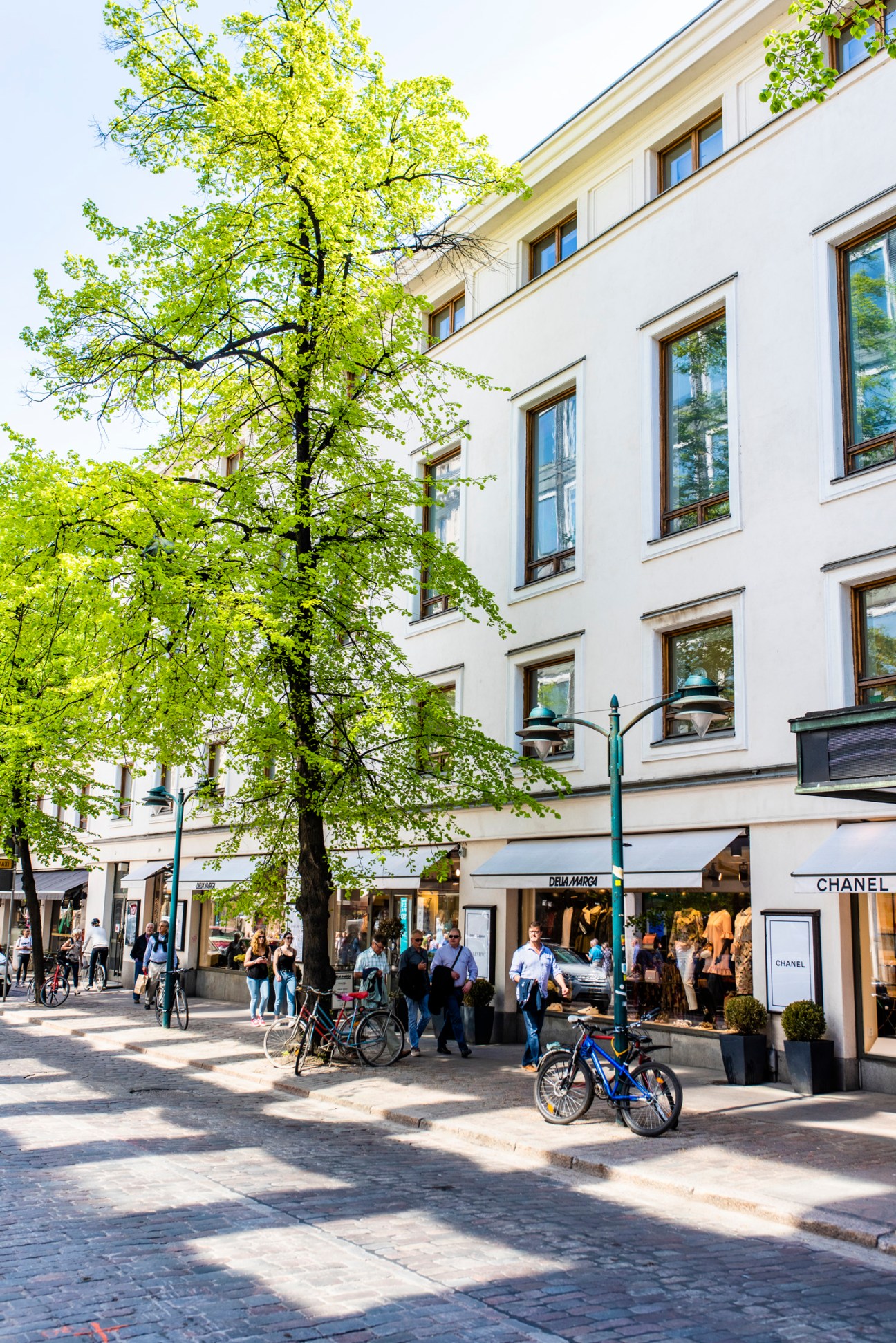 Pedestrians and cyclists on a tree-lined shopping street with white buildings and storefronts including Chanel.