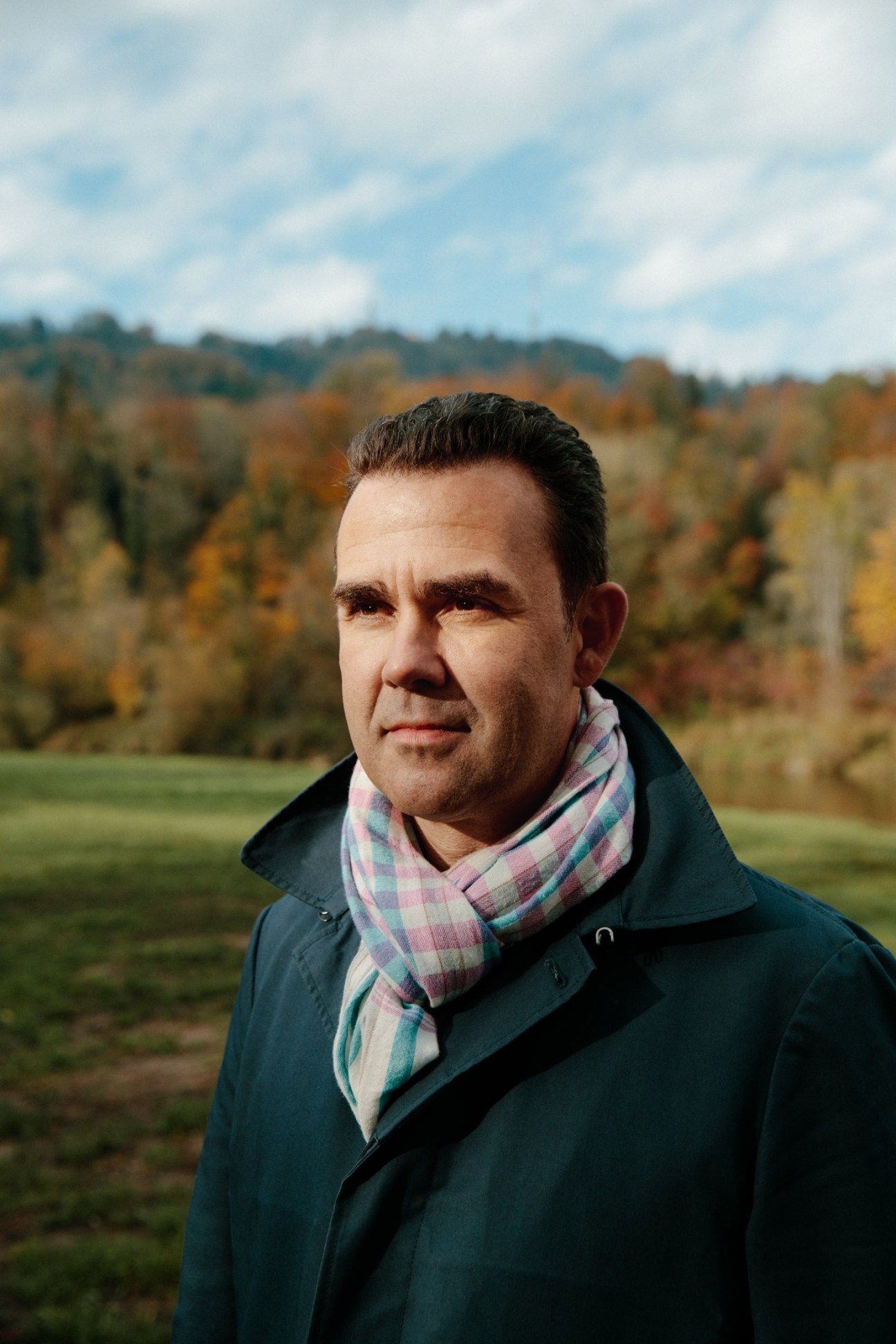 Stephan Ensmann stands outdoors in autumn landscape wearing dark jacket and plaid scarf.