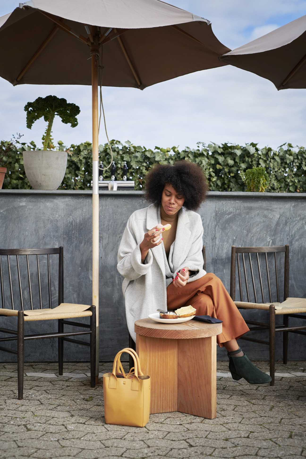 Woman in cream blazer and tan trousers sitting under dark umbrellas at outdoor table with yellow bag and plate of food