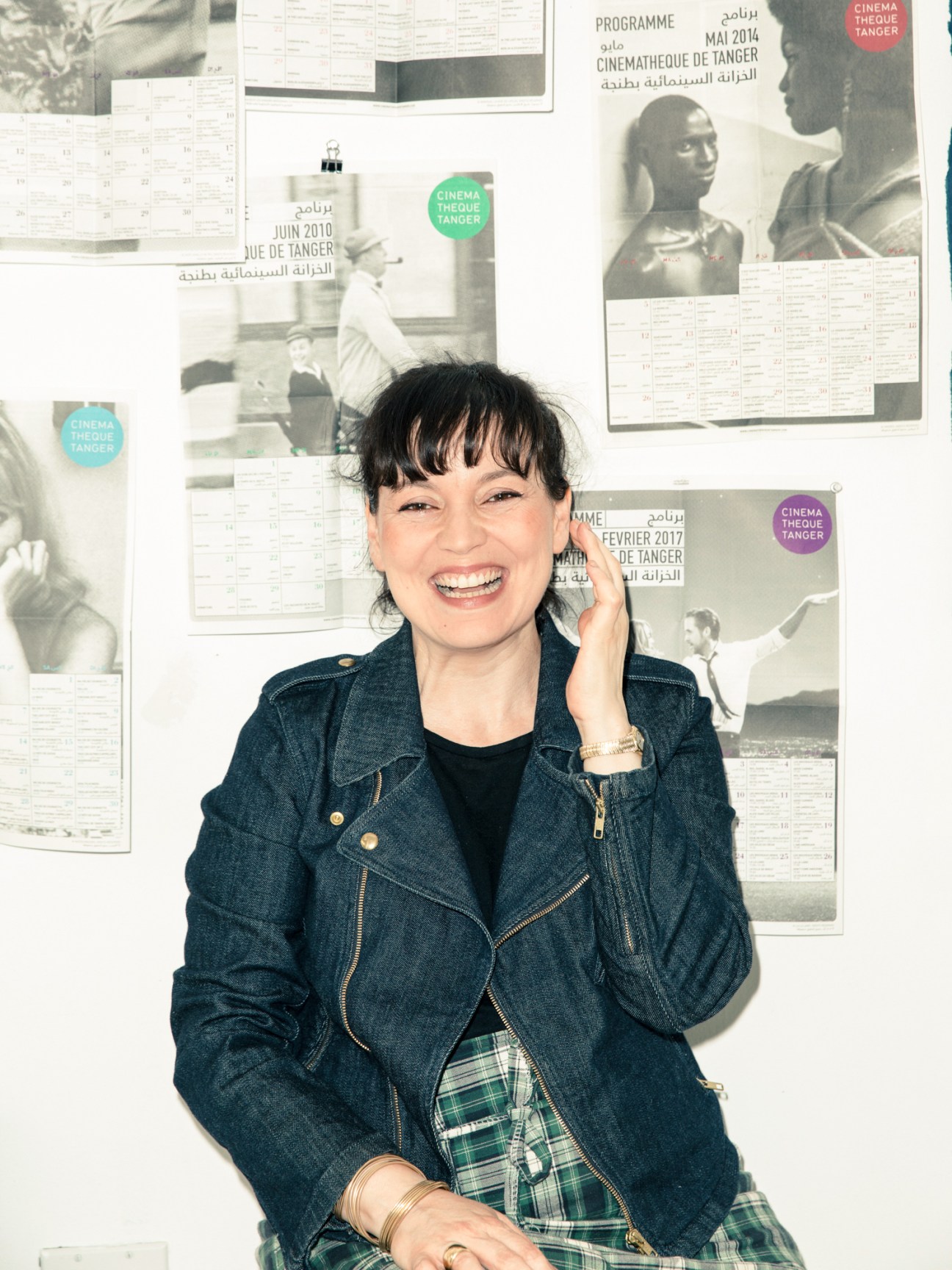 Yto Barrada smiling in front of Cinémathèque de Tanger programme posters on a white wall.