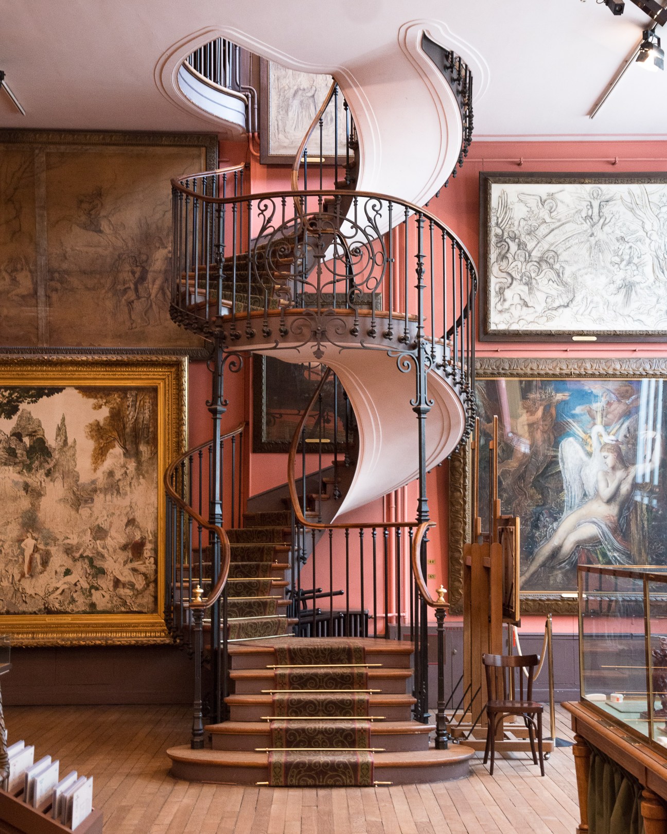 Spiral staircase with ornate ironwork railing and wooden treads at Musée Gustave Moreau in Paris, surrounded by framed artworks.